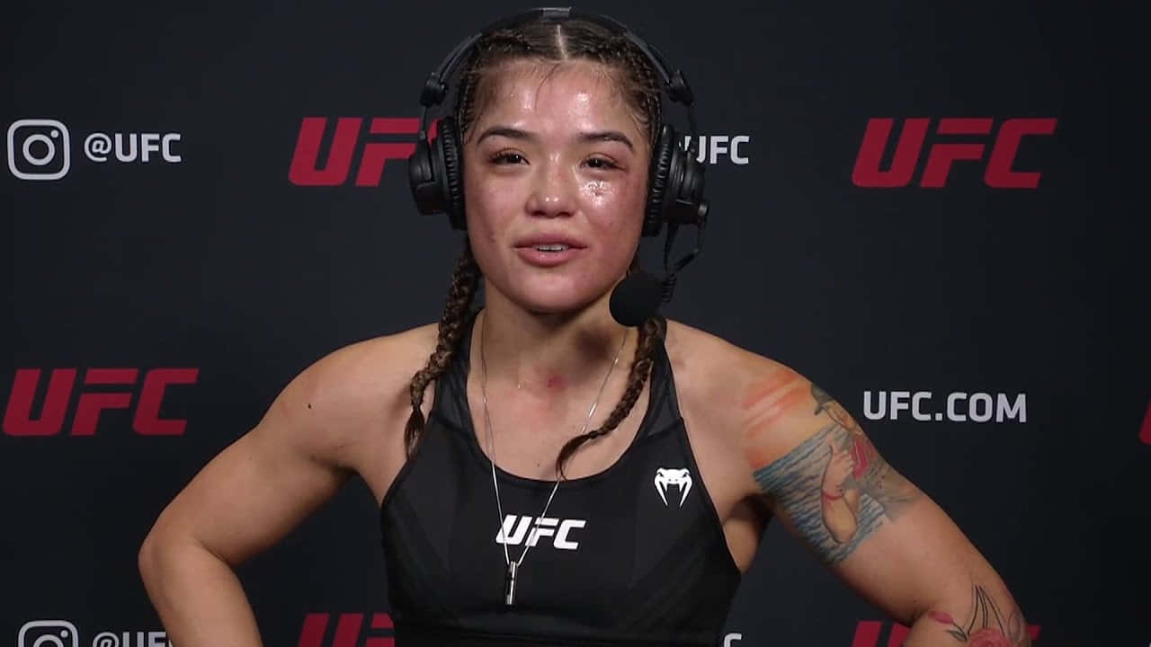  壁紙 — 一位辮子頭髮和紋身的女UFC拳手戴著耳機和黑色背心在記者會上發言。