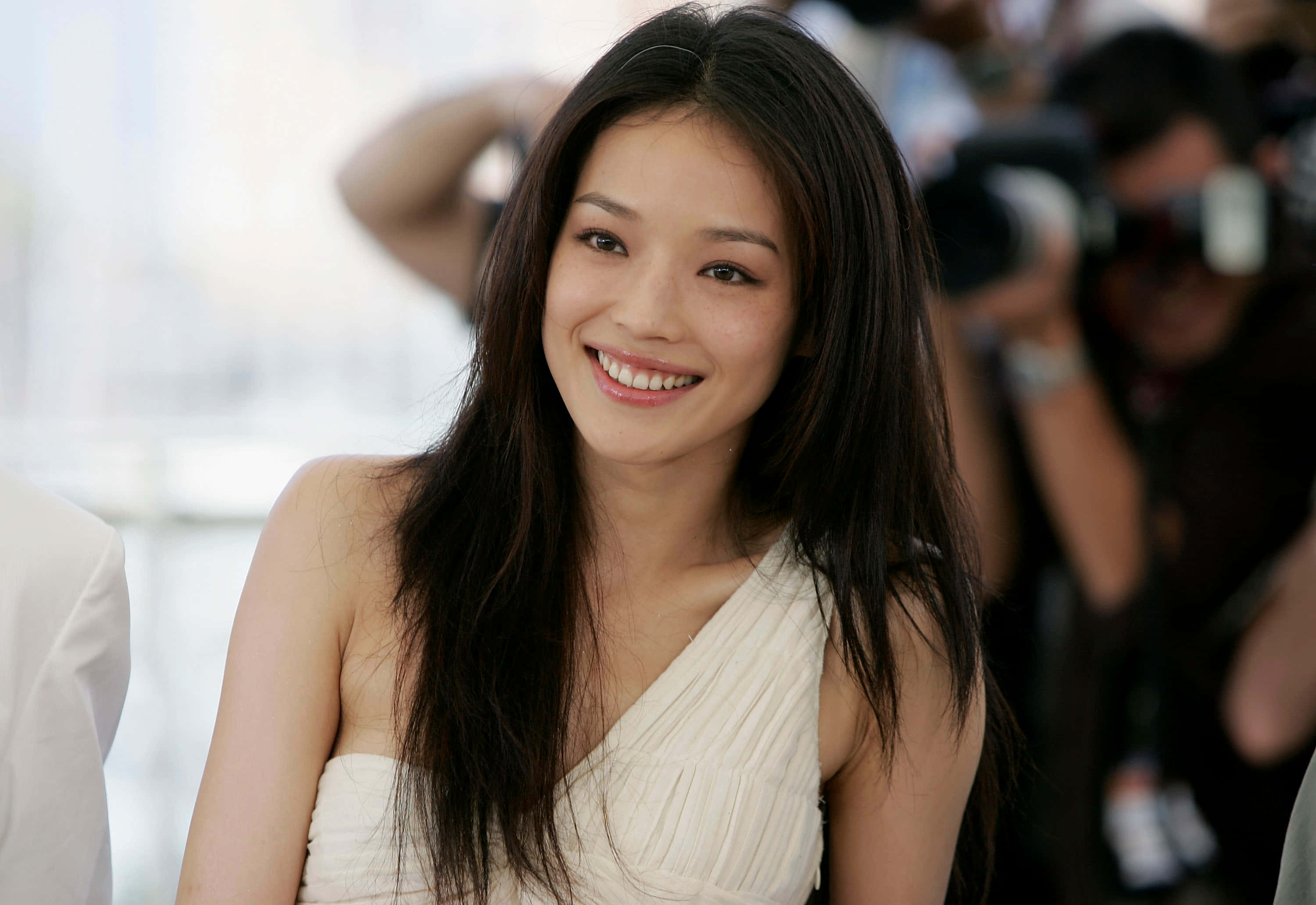 Shu Qi 桌布 壁紙 — 一位長黑髮的燦爛女性穿著白色連衣裙，在繁忙的活動中溫暖微笑，背景有攝影師。