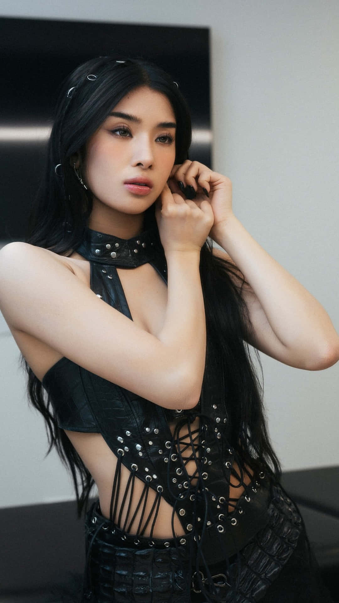  壁紙 — 一位長黑髮的女性穿著帶有鉚釘的黑色皮革服裝，在極簡灰色和黑色背景前將手靠近臉部擺姿勢。