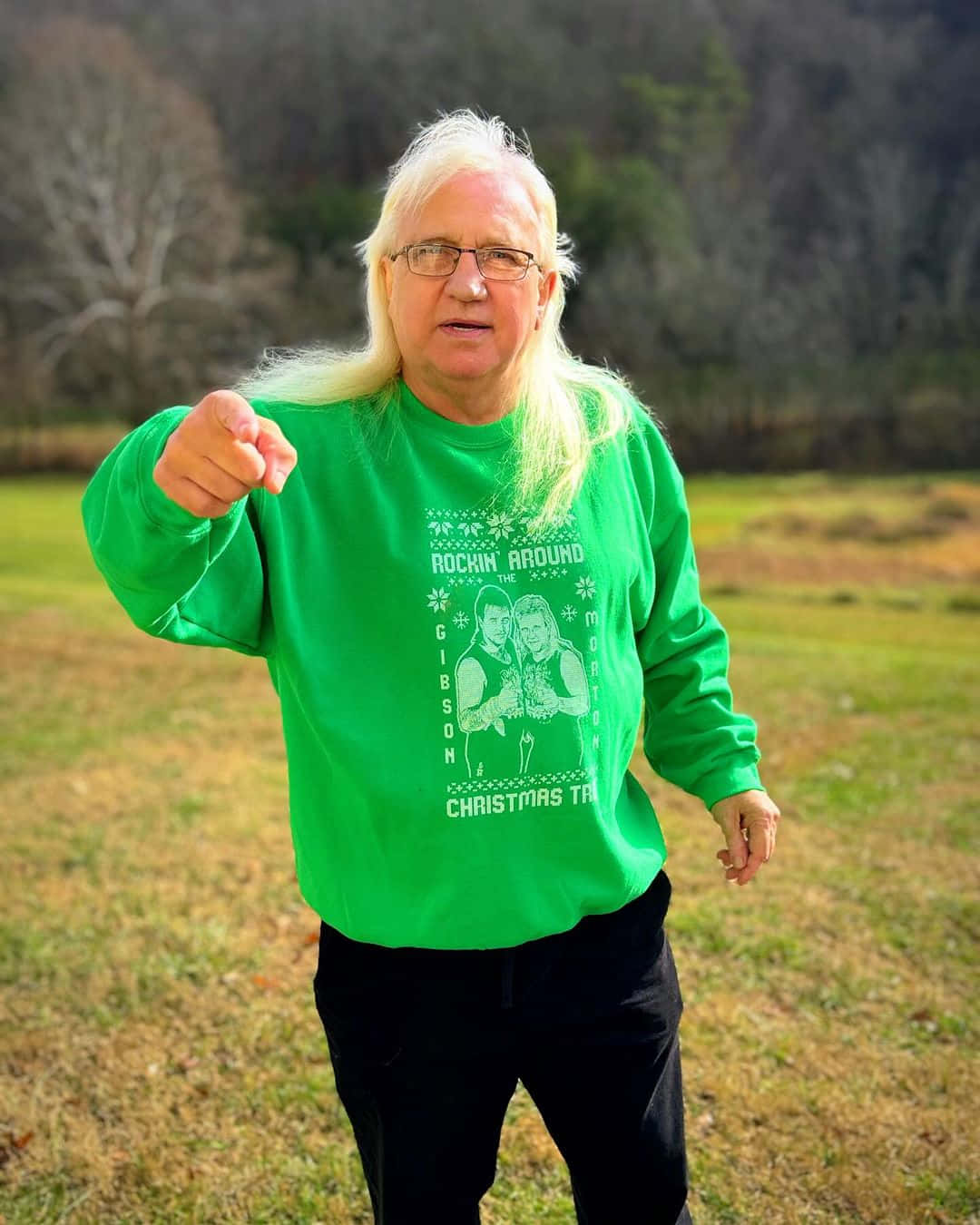  壁紙 — 一位長金髮戴眼鏡的男子，穿著鮮綠色聖誕毛衣，在草地上向前指。