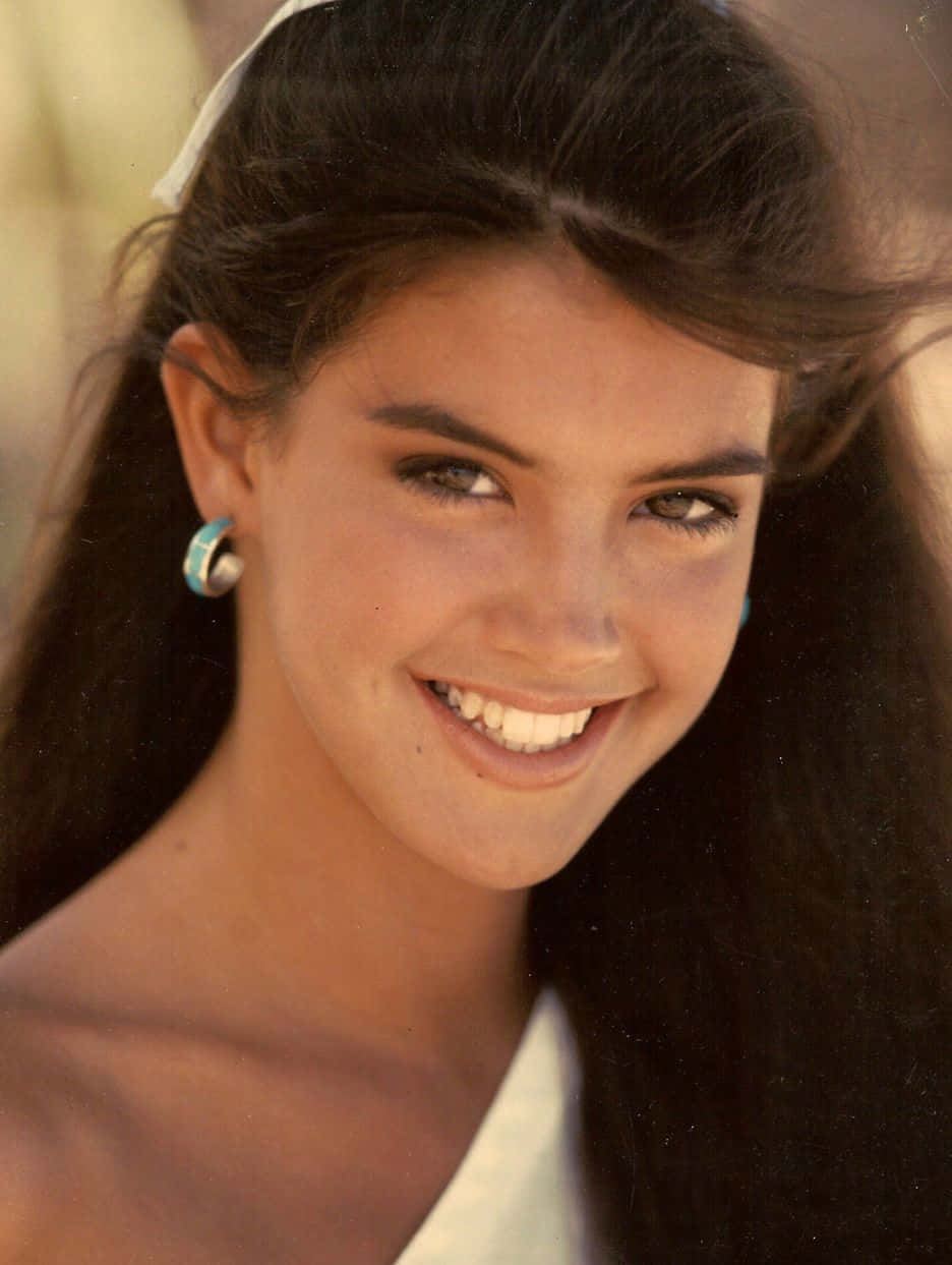 Phoebe Cates 桌布 壁紙 — 一位充滿光彩的年輕女性，深色頭髮，燦爛笑容，在溫暖陽光下直視鏡頭。