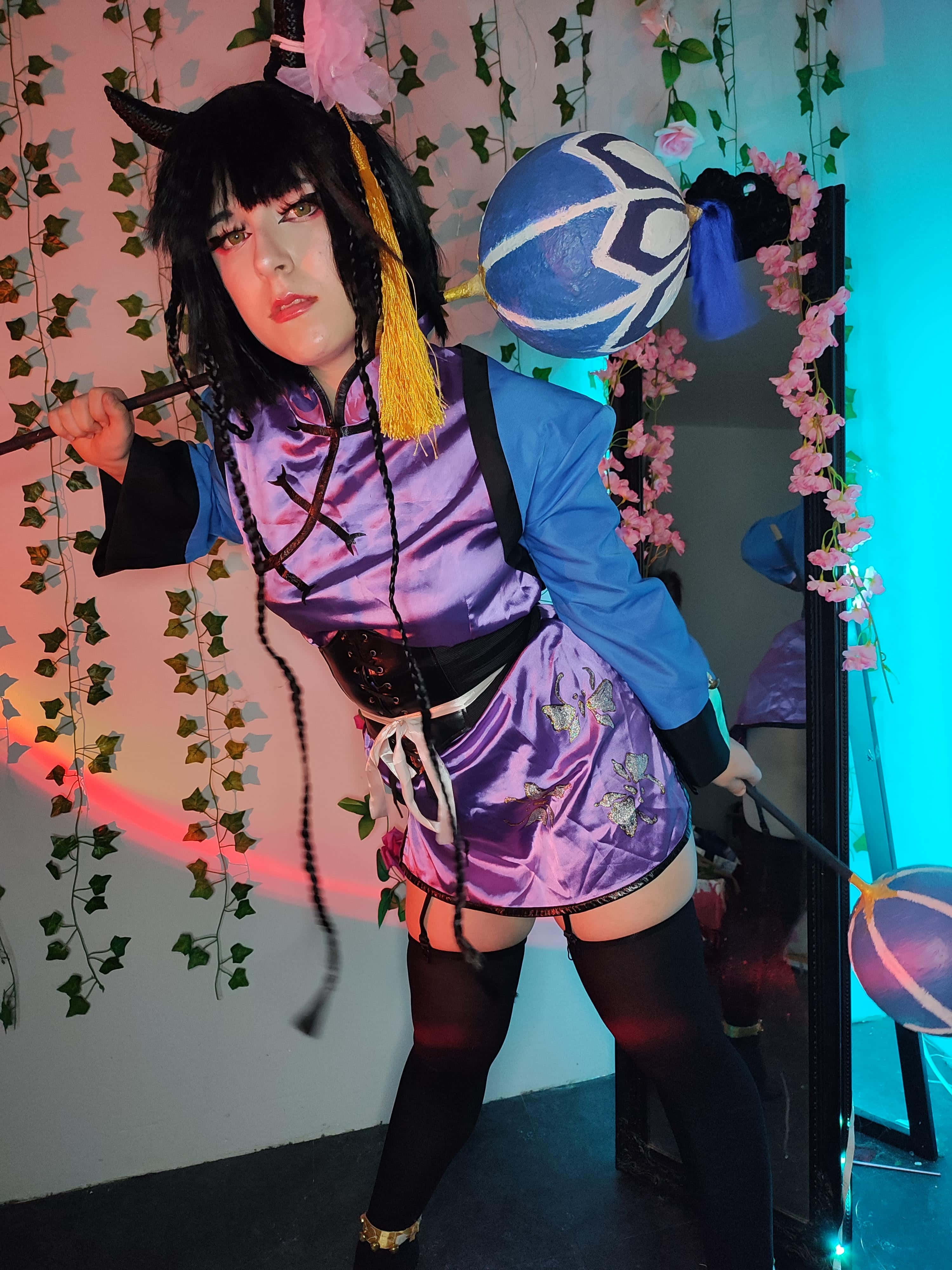 Ran-mao 桌布 壁紙 — 一位穿著紫色和藍色服裝、帶有角和蝴蝶圖案的cosplayer，手持法杖和發光球體，在藤蔓和花朵背景前擺姿勢。