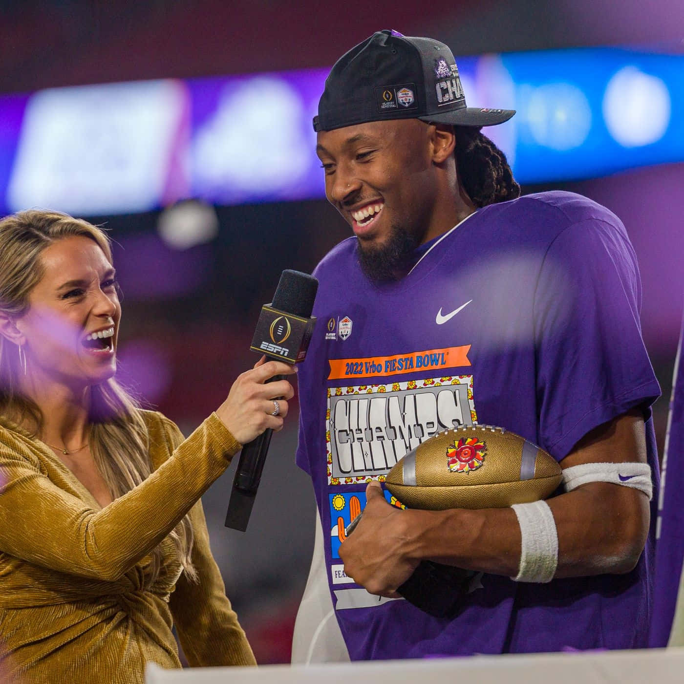  壁紙 — 一位身穿紫色「Champs」球衣的快樂足球員，在燈光明亮的體育館舞台上，一邊捧著獎盃，一邊接受微笑的ESPN記者訪問。
