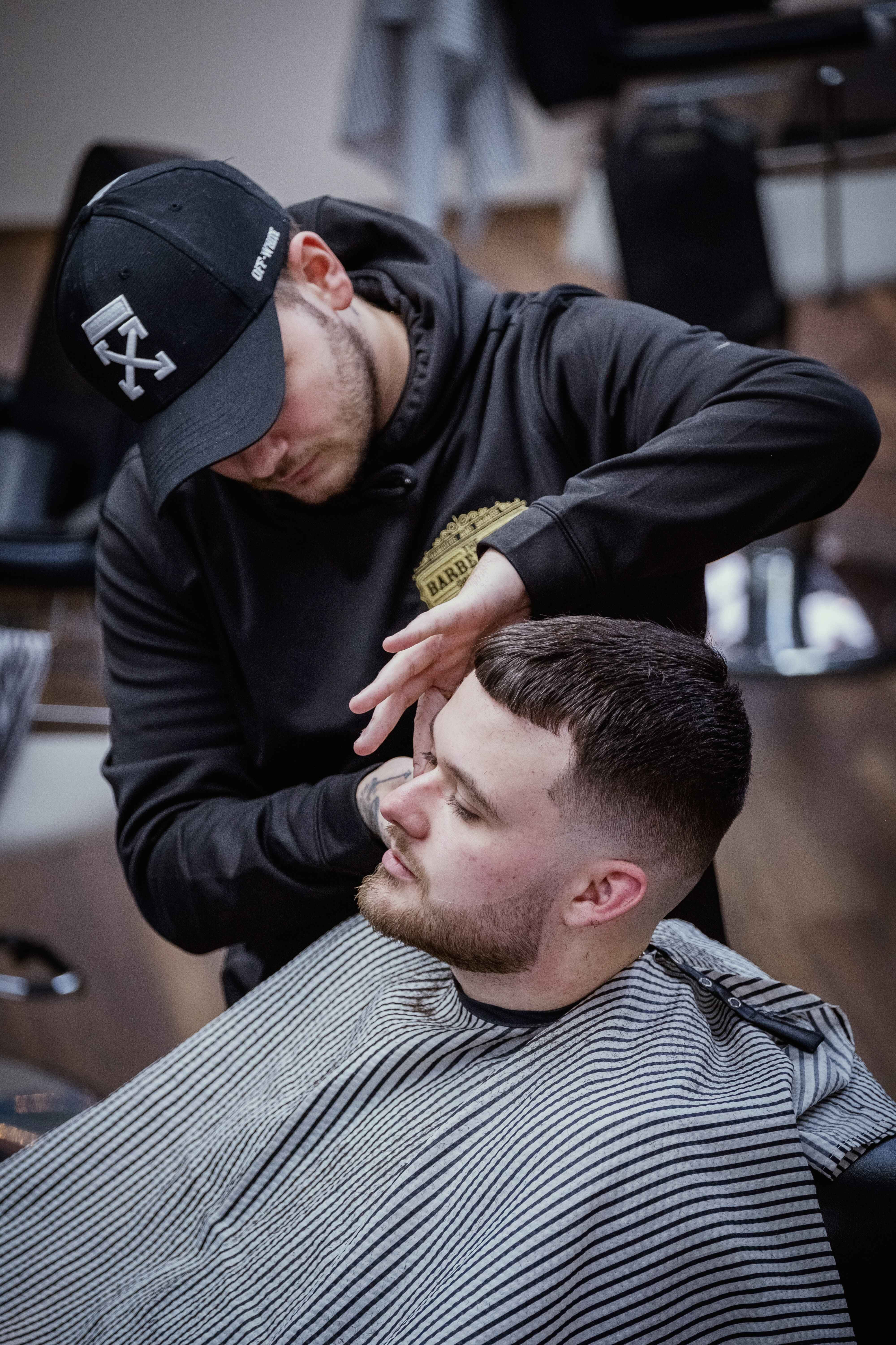 Barber 桌布 壁紙 — 一位戴黑色鴨舌帽和連帽衫的理髮師，在現代理髮店中為顧客小心地整理頭髮，顧客披著條紋圍布。