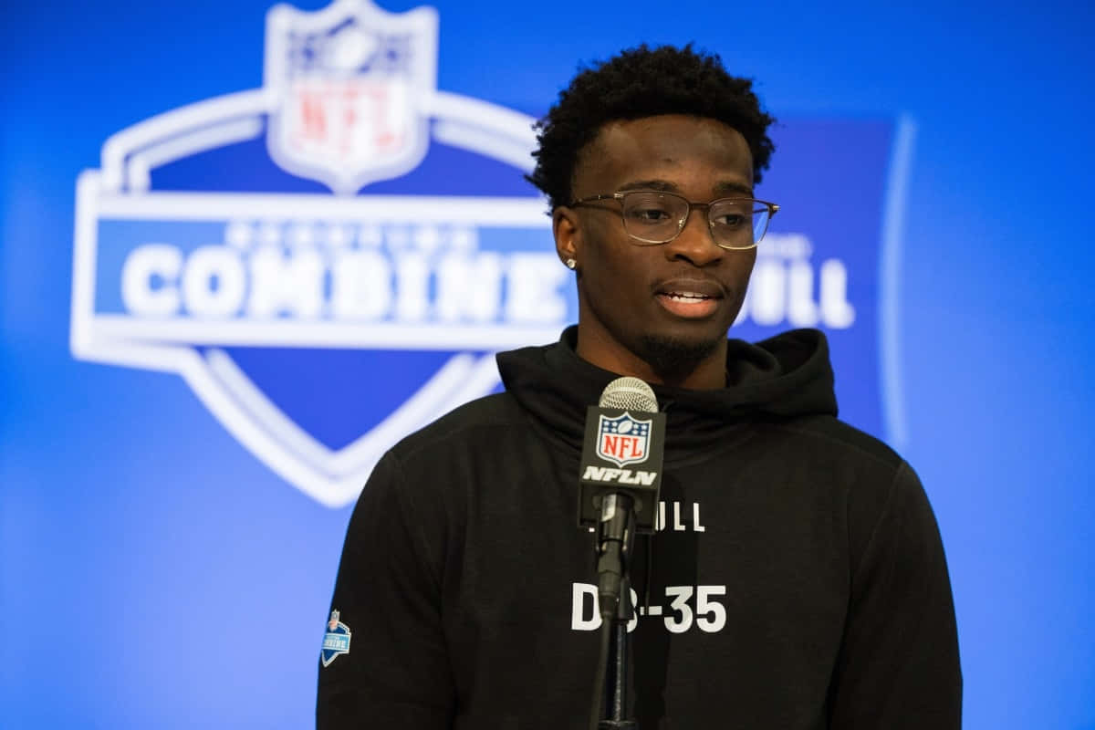  壁紙 — 一位戴眼鏡、穿黑色連帽衫的年輕黑人美式足球員在NFL選秀聯合試訓記者會上對著NFL麥克風發言。