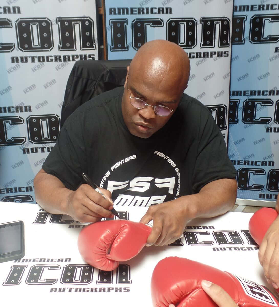 壁紙 — 一位戴眼鏡的光頭男子穿著黑色MMA T恤，在擺有紅色拳擊手套和品牌背景的桌子上簽名。