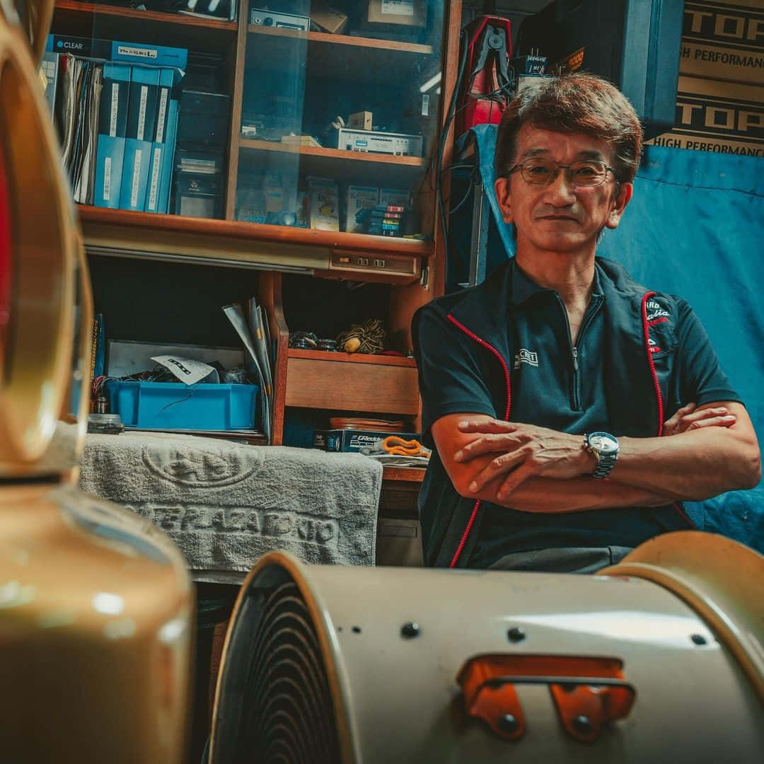  壁紙 — 一位戴眼鏡的長者自信地站在雜亂的車庫工作室中，周圍是工具和汽車零件。