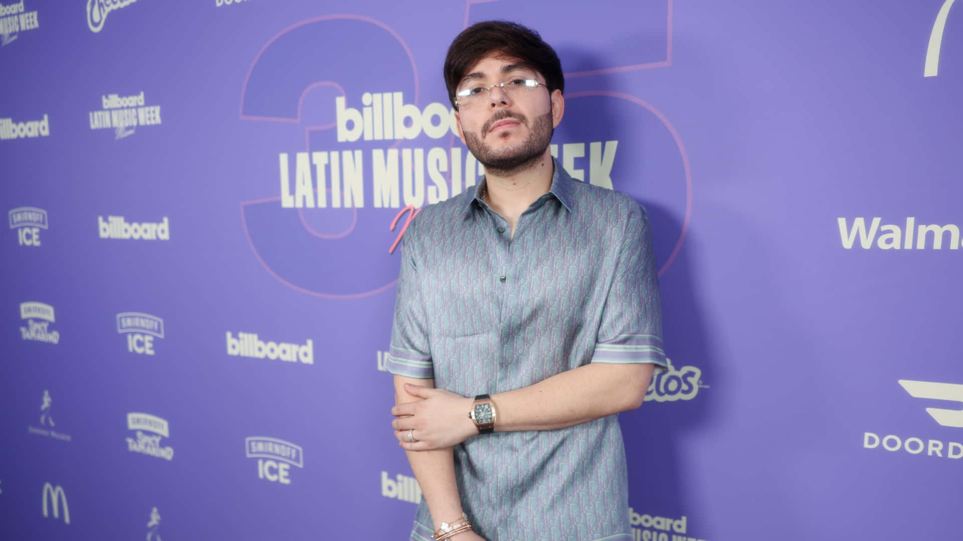  壁紙 — 一位戴眼鏡、留鬍鬚的男子自信地站在帶有Billboard Latin Music Week標誌的紫色活動背景前。