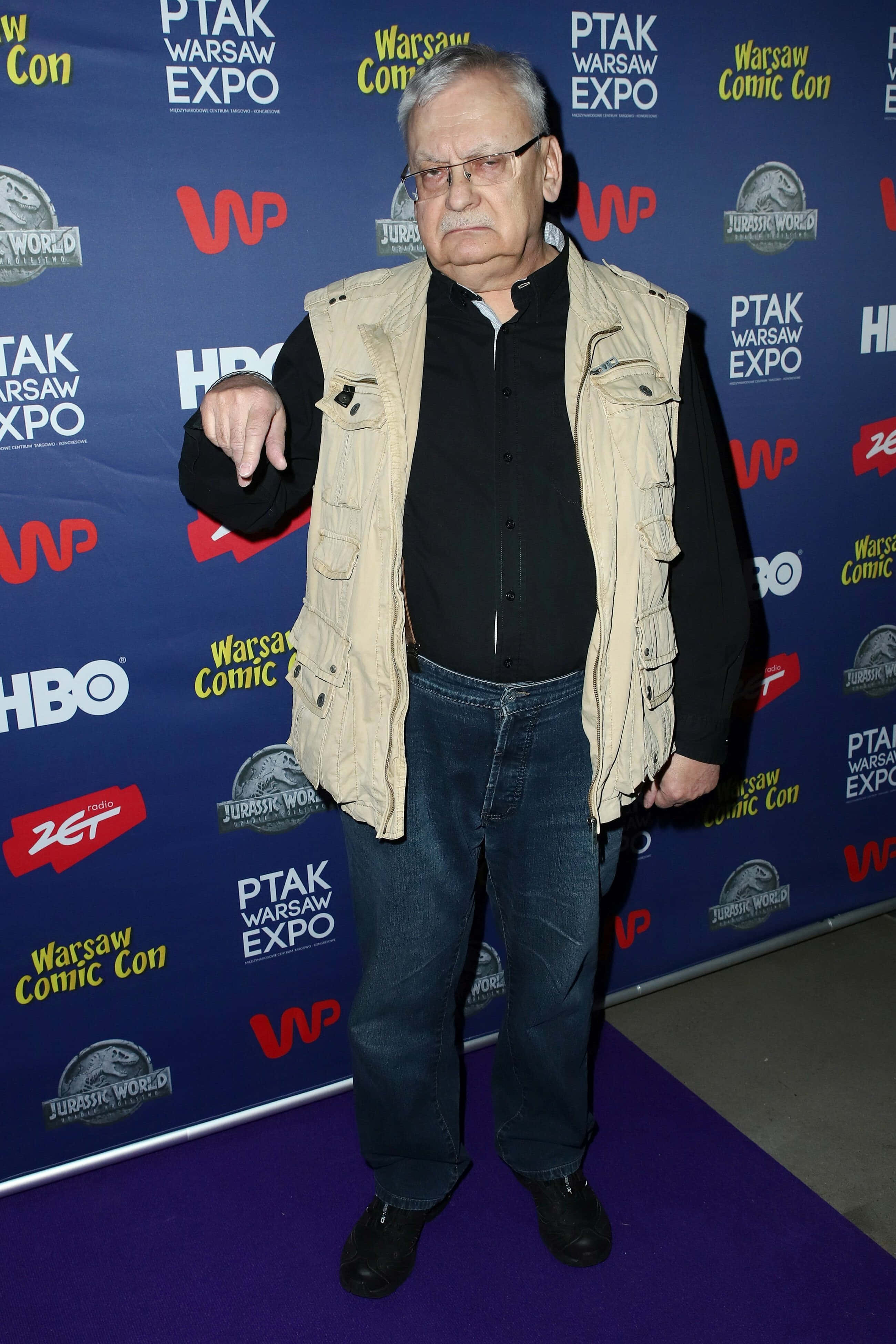 Andrzej Sapkowski 桌布 壁紙 — 一位戴眼鏡、身穿米色背心的年長男士，站在有品牌背景的漫畫展活動的紫色地毯上。
