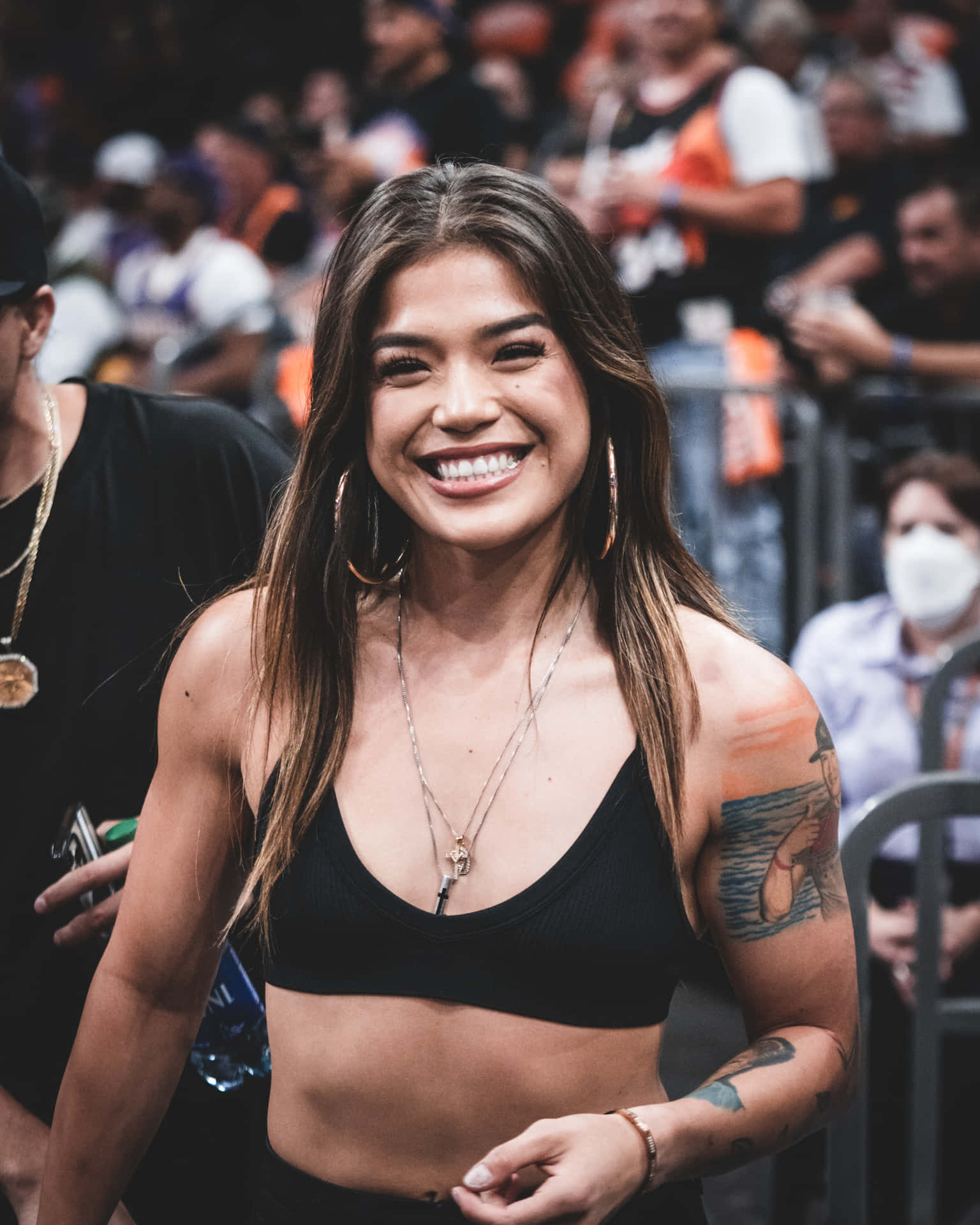  壁紙 — 一位帶有紋身和圓環耳環的微笑女性，在擠滿人的體育場館中對著鏡頭閃耀。