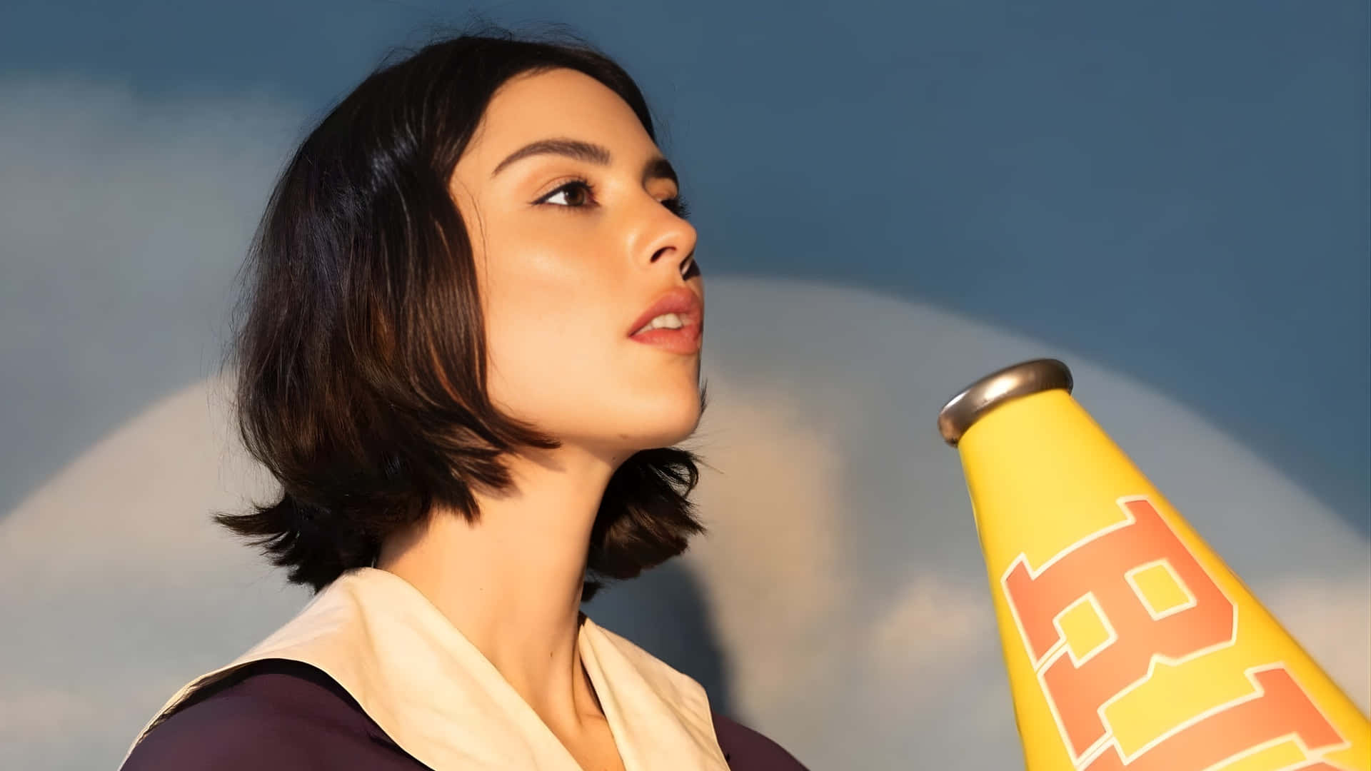  壁紙 — 一位短黑髮的年輕女性在柔和的藍天背景下拿著一個黃色擴音器。