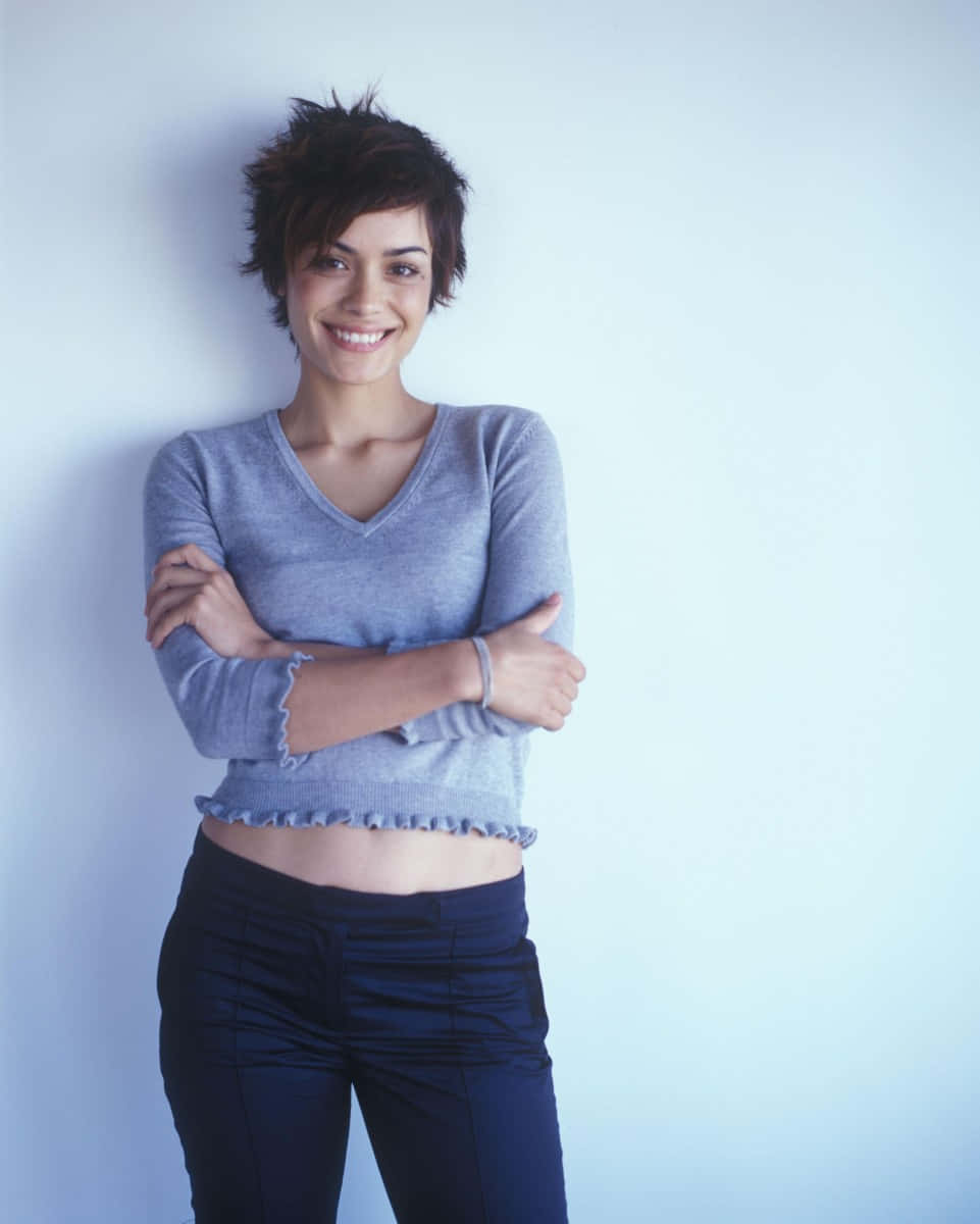 Shannyn Sossamon 桌布 壁紙 — 一位短黑髮微笑的女子靠著淺藍色牆壁，身穿灰色短款毛衣和深色褲子。