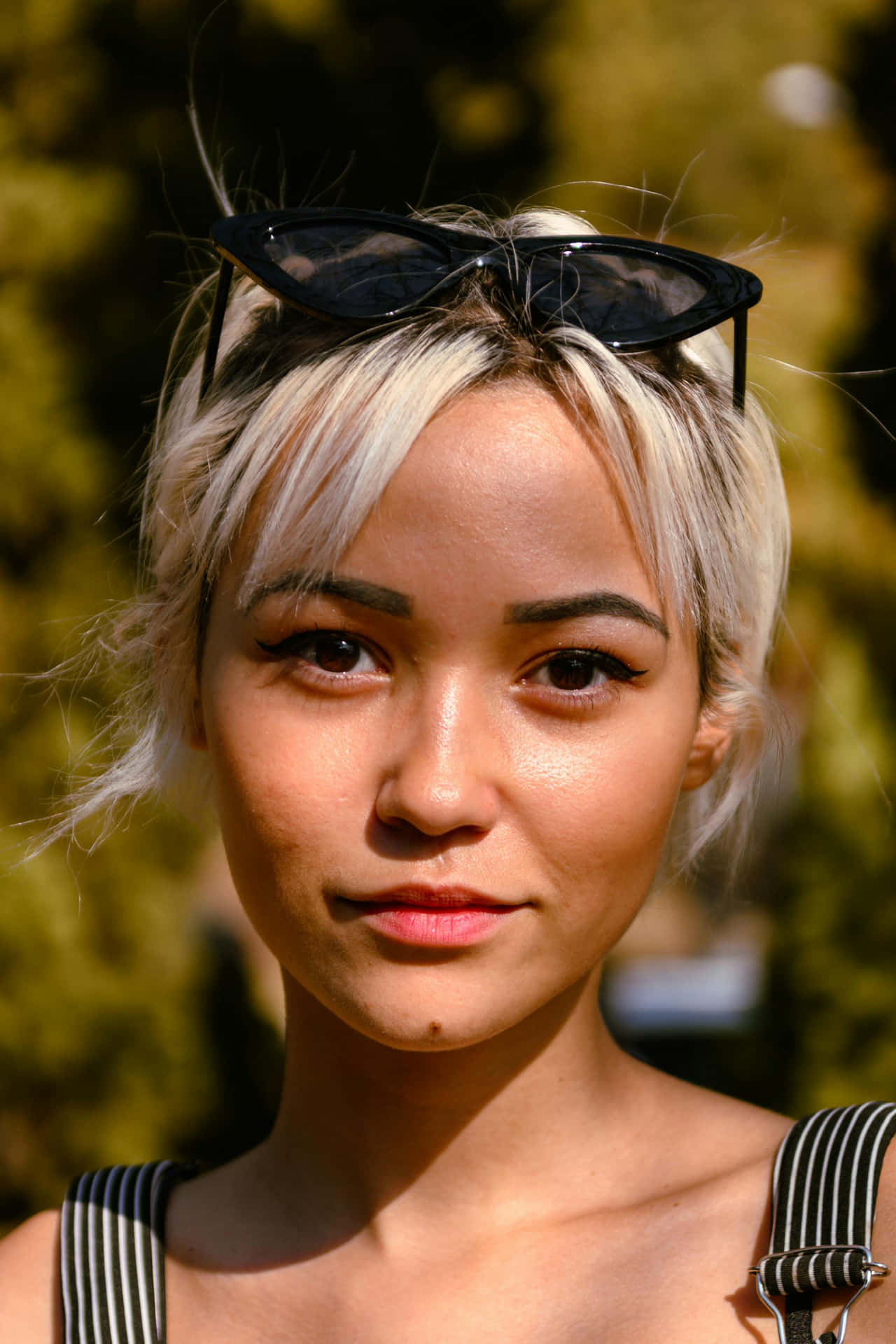  壁紙 — 一位短金髮、頭戴黑色貓眼太陽眼鏡的年輕女性，在陽光明媚的綠色背景下直視鏡頭。
