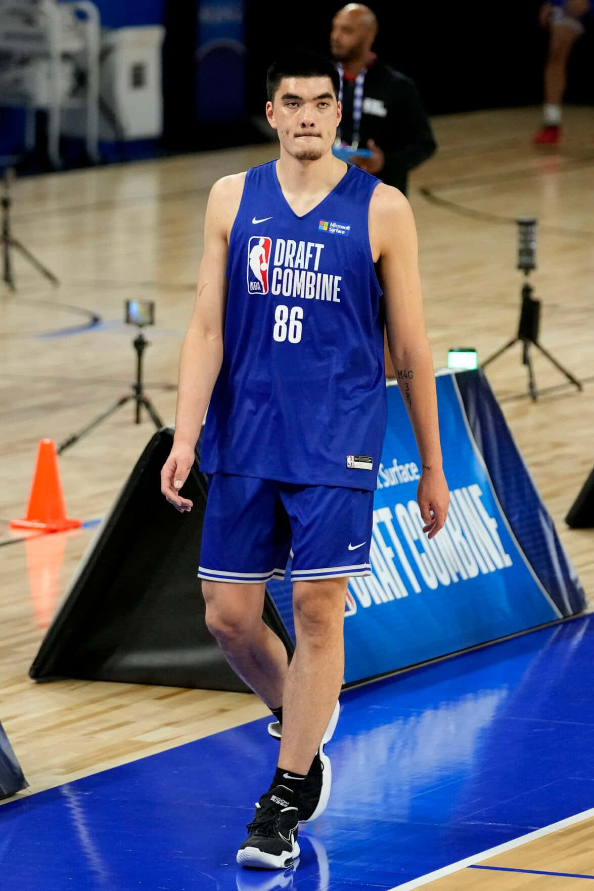  壁紙 — 一位高大的籃球運動員身穿藍色「Draft Combine」球衣，自信地走過拋光球場，背景有橙色錐體和相機。