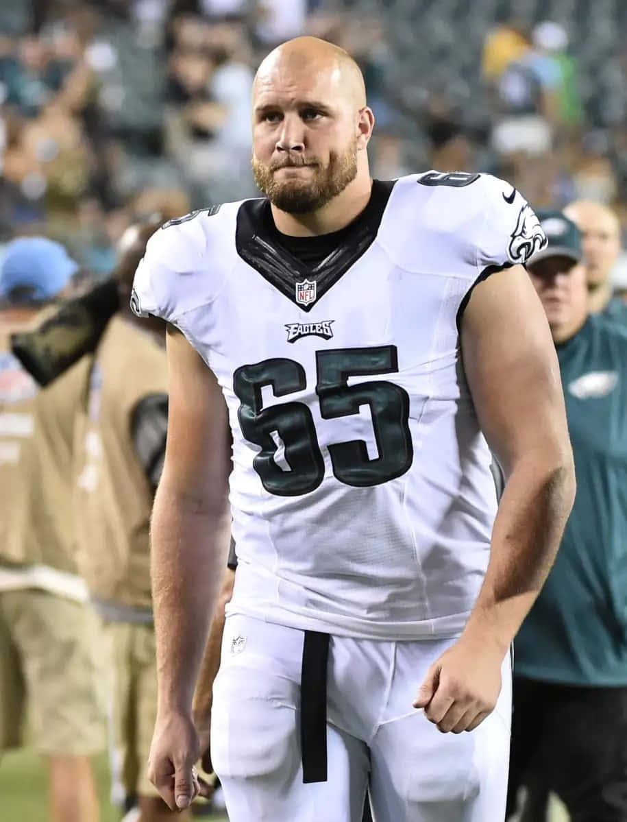  壁紙 — 一位光頭NFL美式足球員，身穿白色Eagles球衣，號碼65，站在球場上，球場燈光照射。