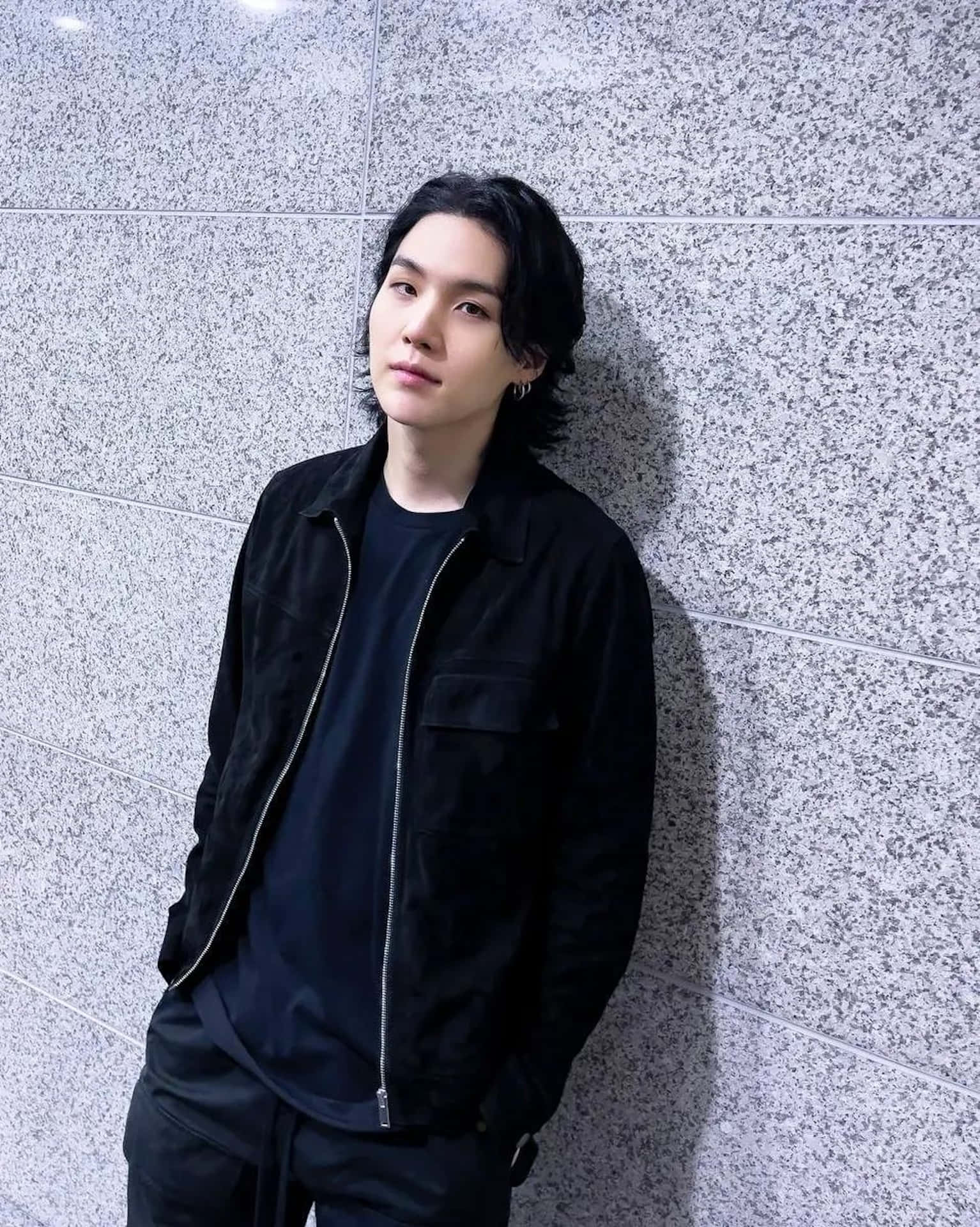  壁紙 — 一位黑髮年輕男子身穿全黑衣服，隨意地靠在一面灰色斑點瓷磚牆上。