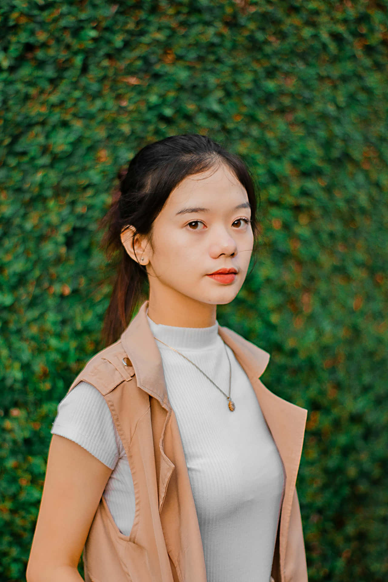  壁紙 — 一位黑髮、塗橙色唇膏的年輕女性站在茂密的綠色常春藤牆前，身穿米色背心內搭白色高領衫。