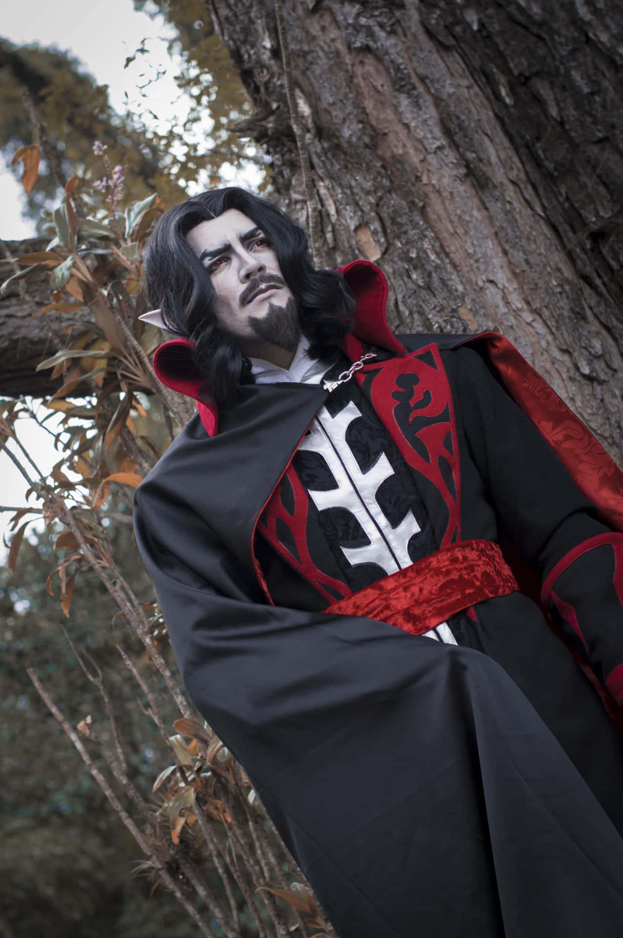  壁紙 — 一位化著深色妝容、戴著尖耳朵的cosplayer，身穿帶有銀色十字繡的黑色和紅色哥德式長袍，靠在一棵多節的樹上。