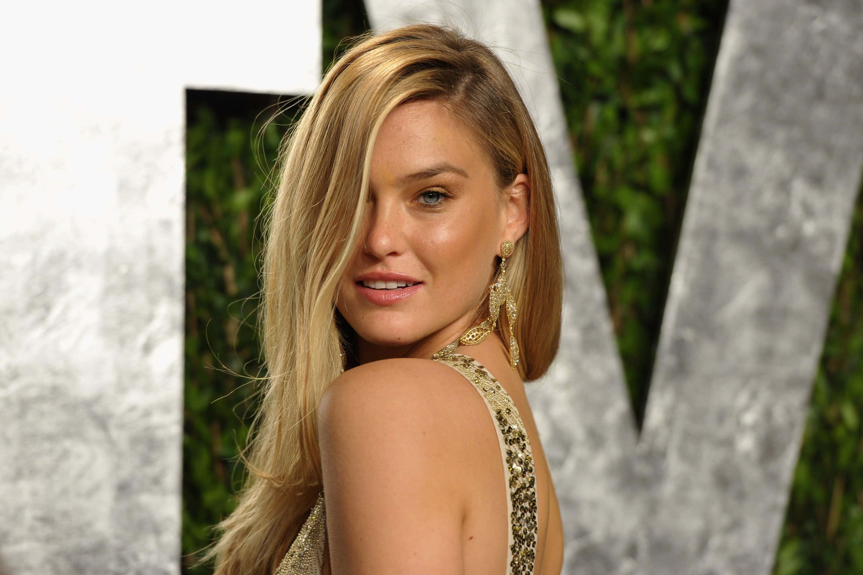Bar Refaeli 桌布 壁紙 — 一位金髮女子身穿金色亮片連衣裙，在綠色樹籬背景襯托下，回頭微笑，背景上有大型白色字母。