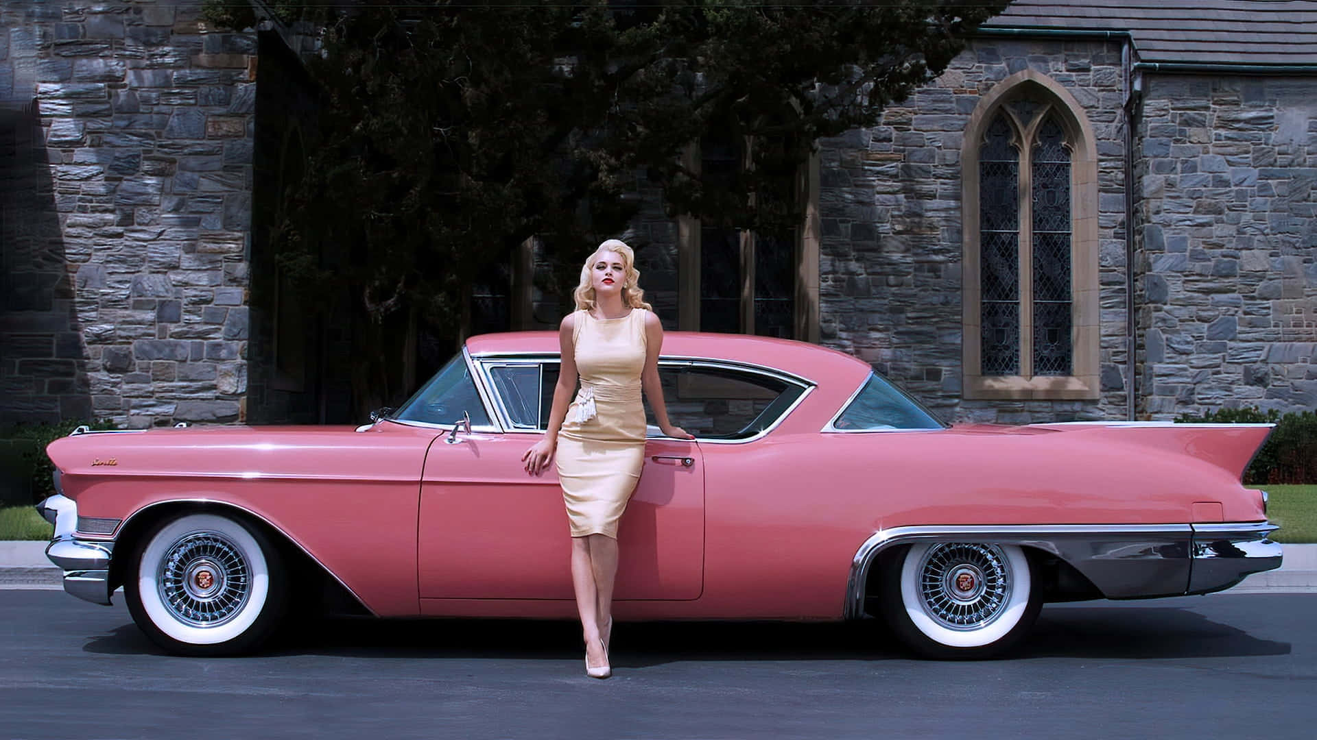 Cadillac Eldorado 桌布 壁紙 — 一位金髮女子身穿米色連衣裙，靠在一輛停放在石砌教堂前的復古粉紅色凱迪拉克上，教堂有拱形窗戶。