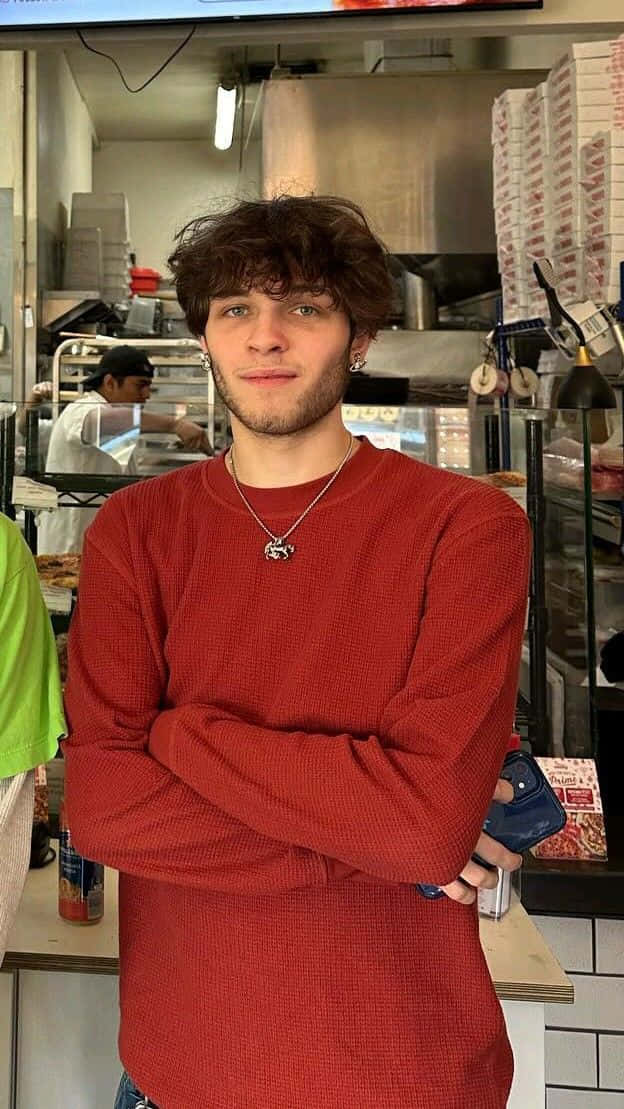  壁紙 — 一位捲髮、身穿紅色毛衣的年輕男子，在繁忙的薄餅店廚房裡自信地交叉雙臂站立。