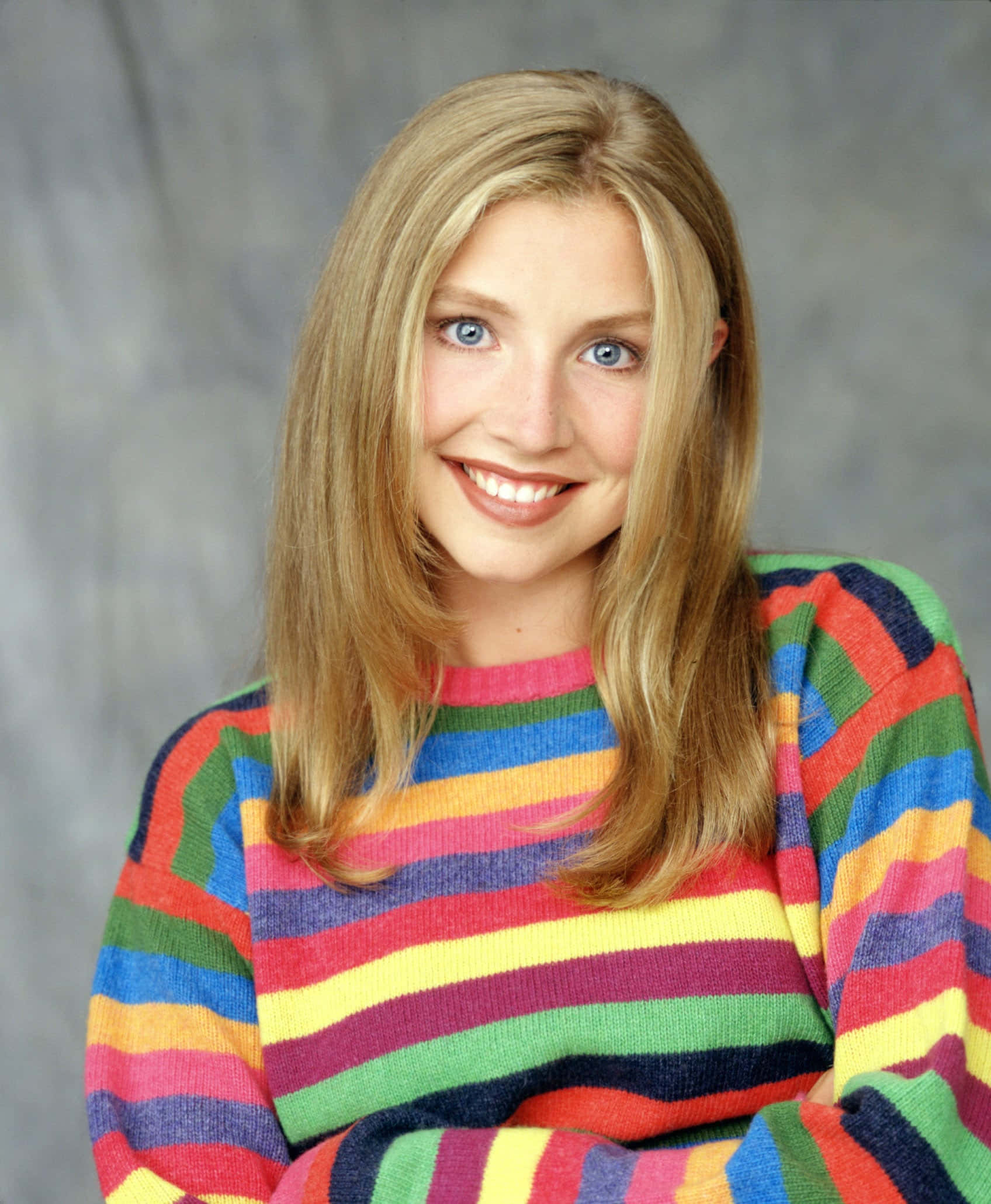 Sarah Chalke 桌布 壁紙 — 一位開朗的年輕女性，金髮碧眼，穿著充滿活力的彩虹條紋毛衣，在灰色中性背景前溫暖地微笑。