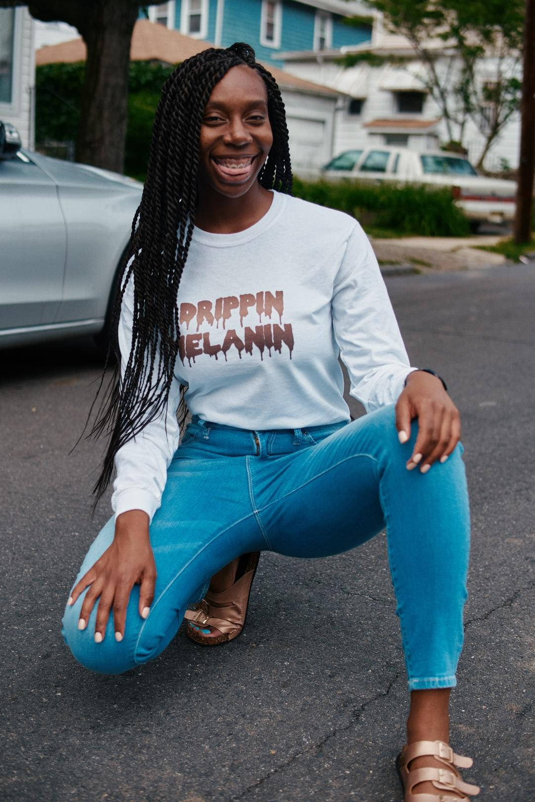 女士蹲在街上，穿著 'Prippin Melanin' 襯衫和藍色牛仔褲。