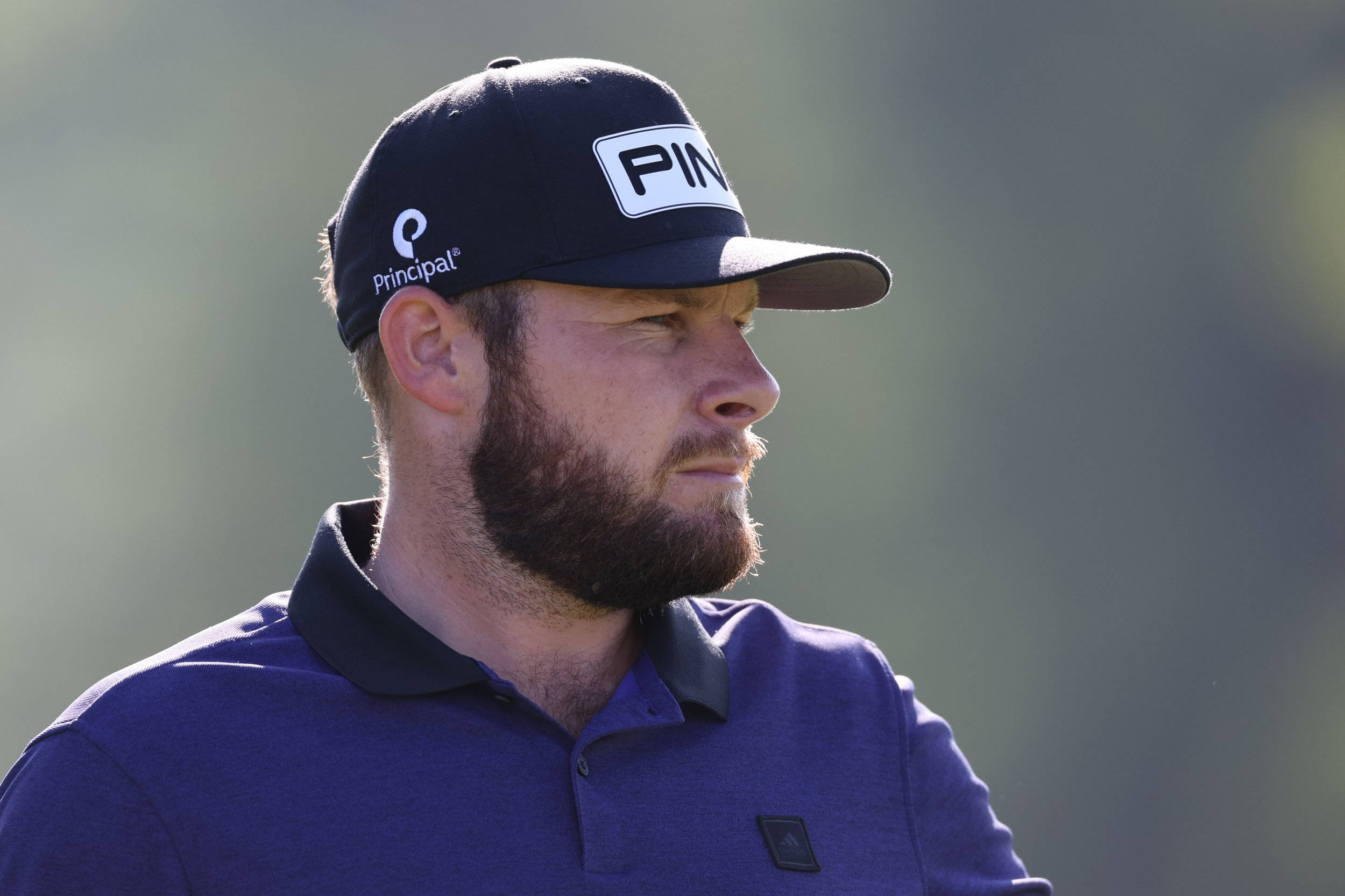 Tyrrell Hatton 桌布 壁紙 — 一位留鬍鬚、專注的高爾夫球手，戴著海軍藍帽子和Polo衫，背景是模糊的綠色。