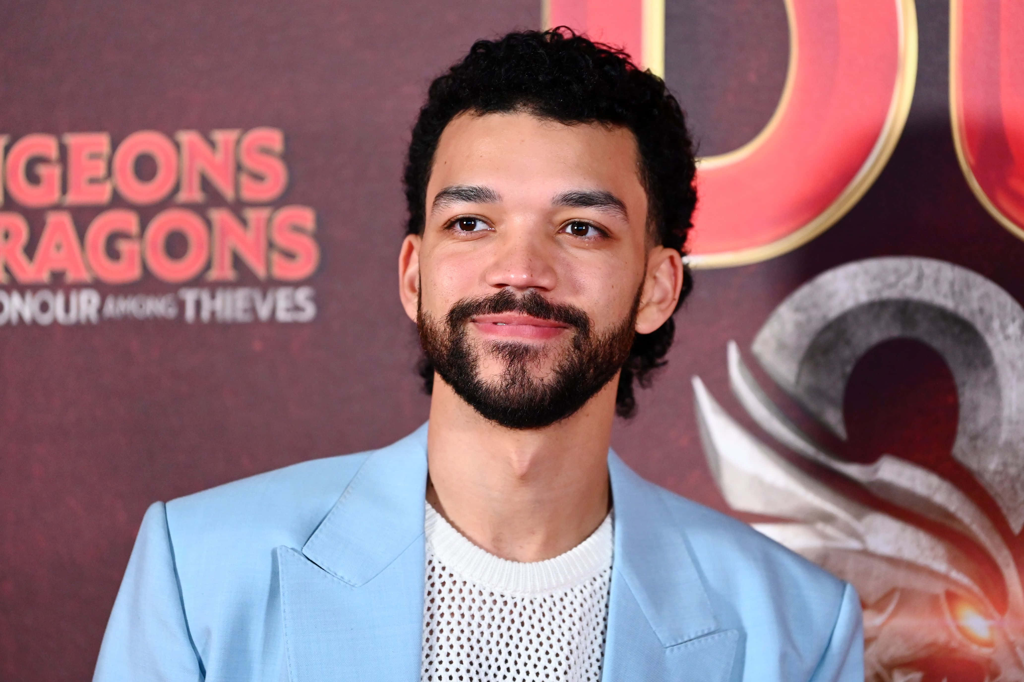 Justice Smith 桌布 壁紙 — 一位留鬍子、捲髮的男子穿著淺藍色西裝外套，在龍與地下城活動背景中微笑。