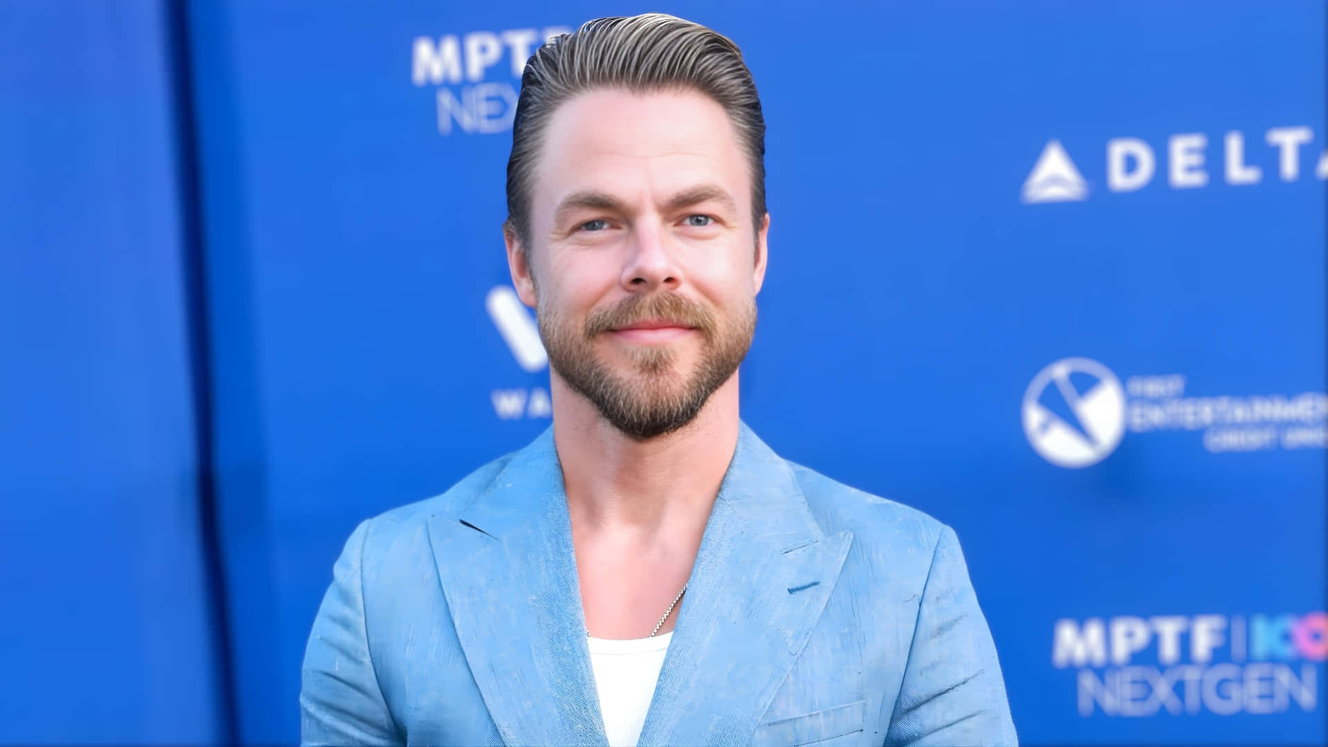  壁紙 — 一位留鬍子、頭髮造型時尚的男子，身穿淺藍色西裝，在品牌藍色背景前微笑。