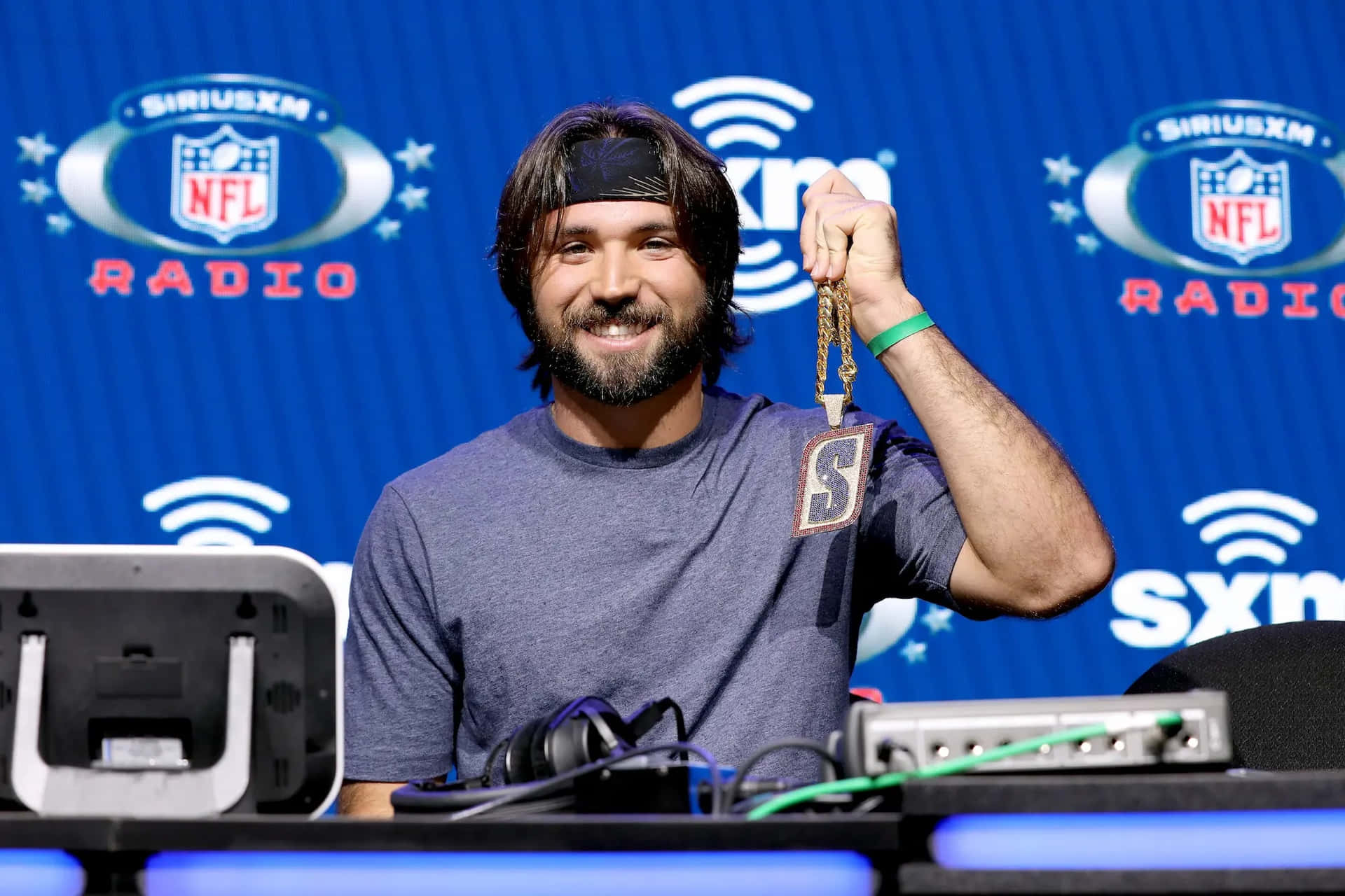  壁紙 — 一位留鬍鬚的 smiling 男士在藍色 SiriusXM NFL Radio 背景前舉起一個金色 NFL 收音機鎖匙扣。