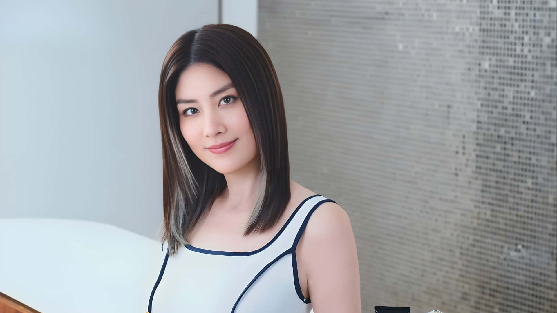  壁紙 — 一位留著及肩深色頭髮的女性，穿著帶有黑色飾邊的白色上衣，在現代瓷磚牆前溫柔微笑。