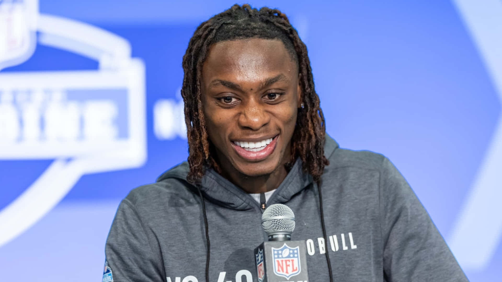  壁紙 — 一位面帶微笑、梳著辮子的NFL球員在記者會上穿著灰色連帽衫對著麥克風說話。