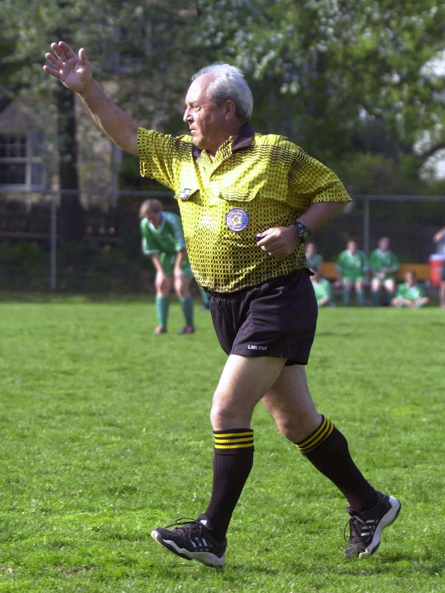 Soccer Referee 桌布 壁紙 — 一位年長男性足球裁判身穿亮黃色球衣，在陽光明媚的草地上果斷地做手勢，背景中有穿著綠色球衣的球員。