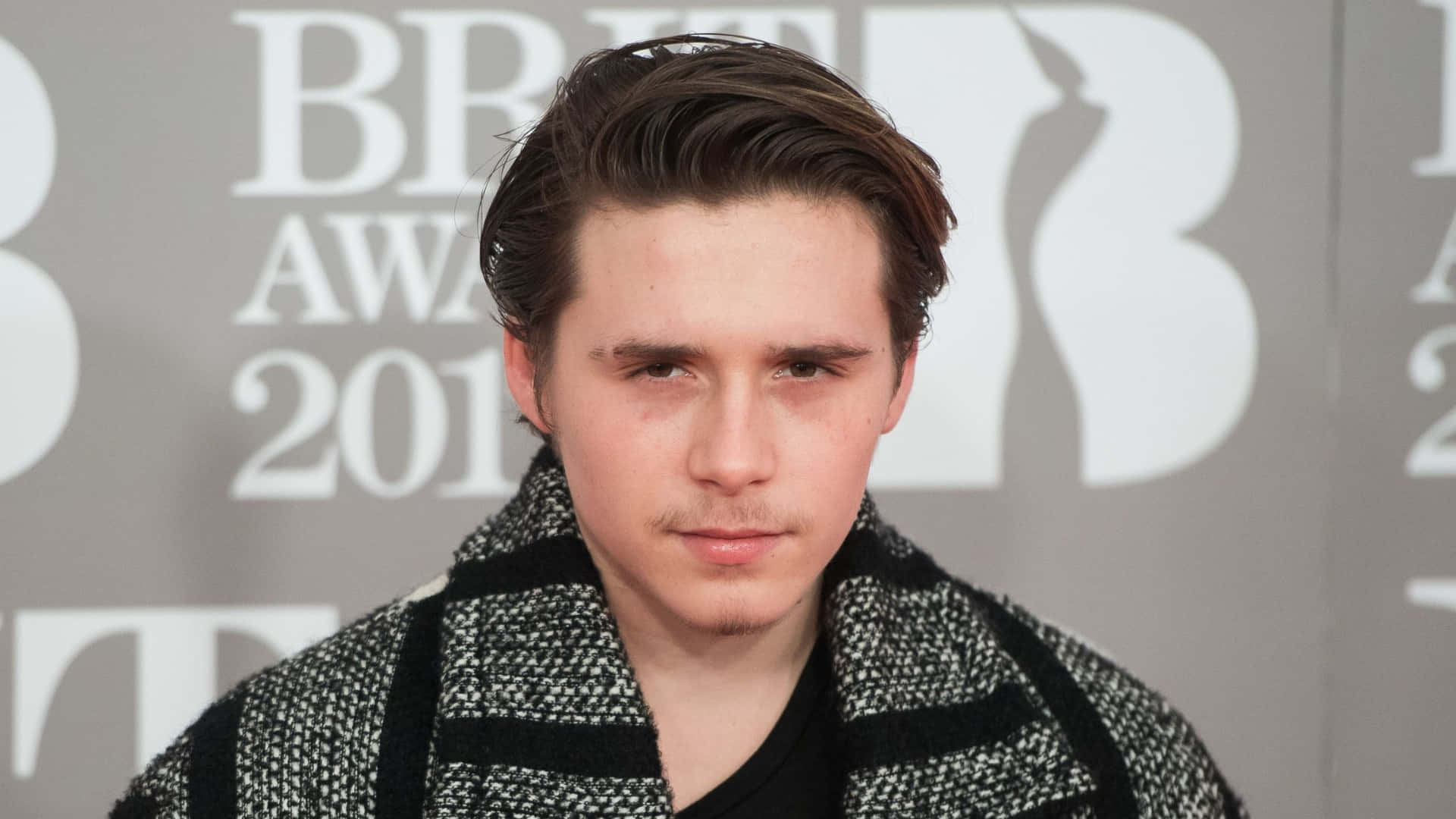  壁紙 — 一位年輕男子，留著時尚的棕色頭髮，戴著圖案圍巾，在灰色BRIT Awards 2013背景前直視鏡頭。