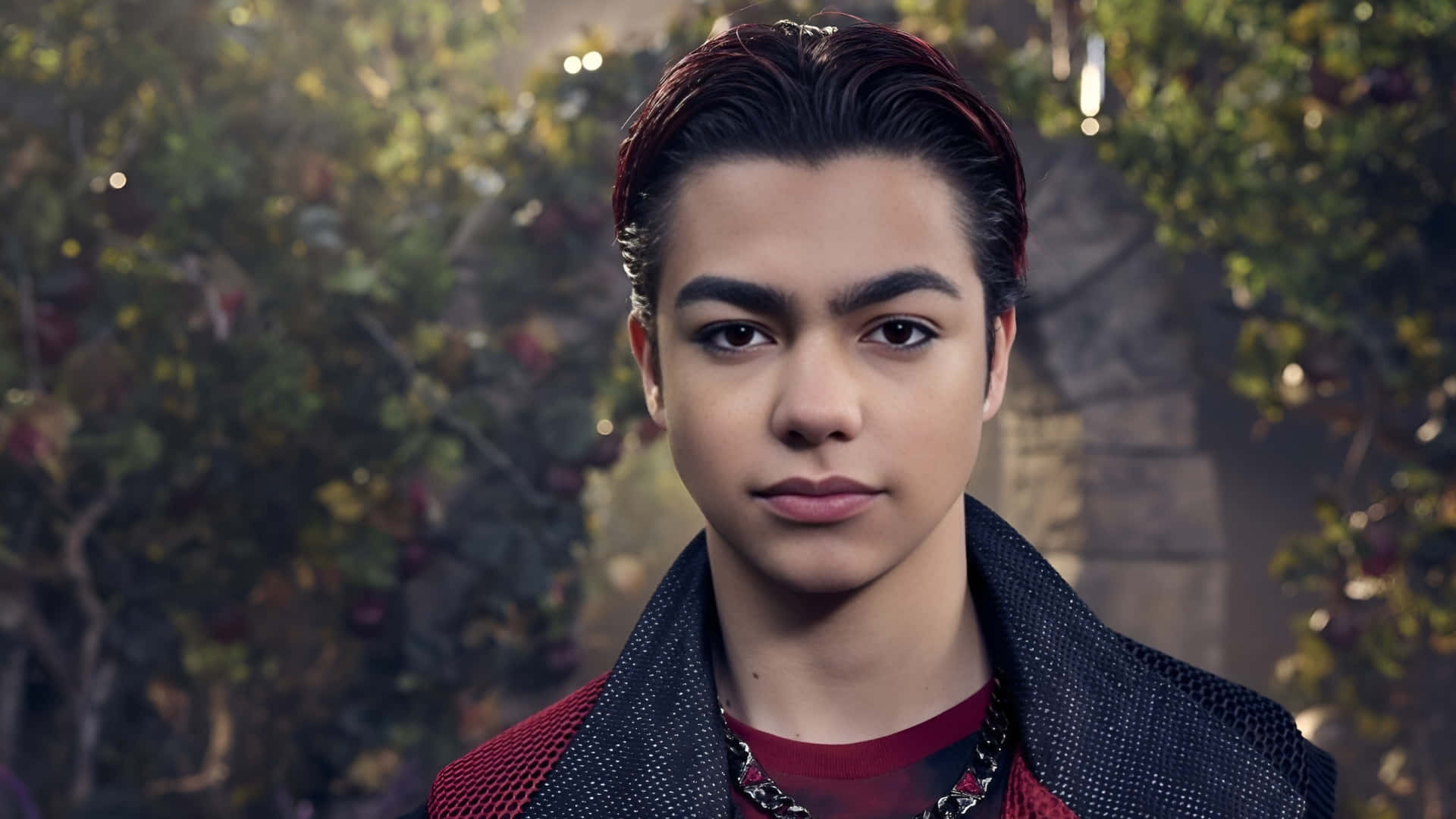  壁紙 — 一位年輕男子，留著造型深色頭髮和紅色挑染，在柔和模糊的花園背景下直視鏡頭。
