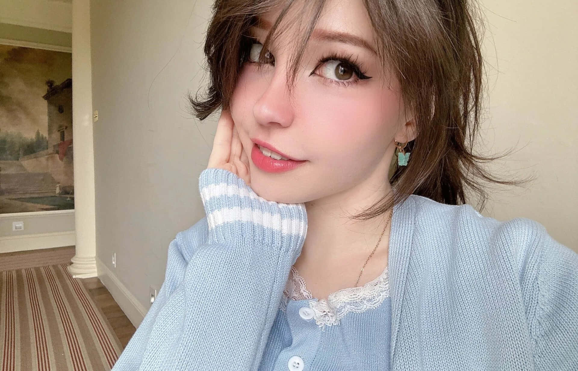 壁紙 — 一位年輕女性化著淡妝，身穿淺藍色開襟毛衣，在室內將手輕輕放在臉頰上擺姿勢。
