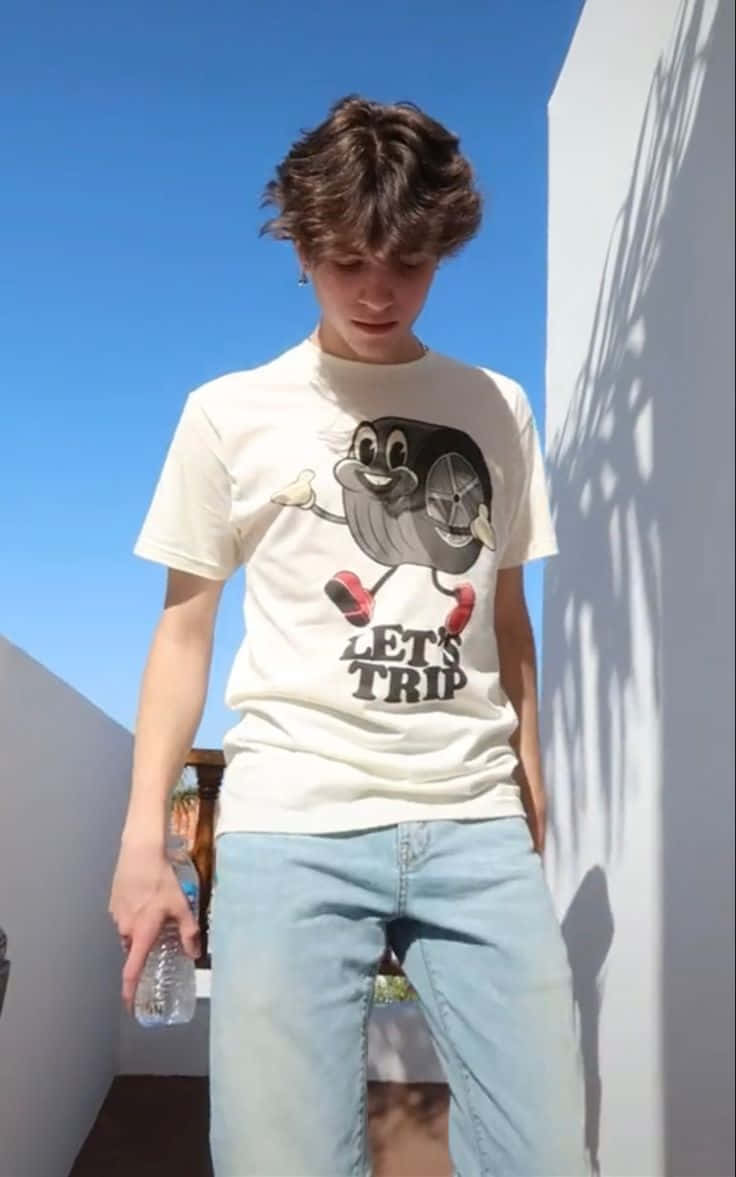  壁紙 — 一位身穿白色圖案T恤和淺色牛仔褲的年輕男子，在晴朗的藍天下站在陽光普照的陽台上。