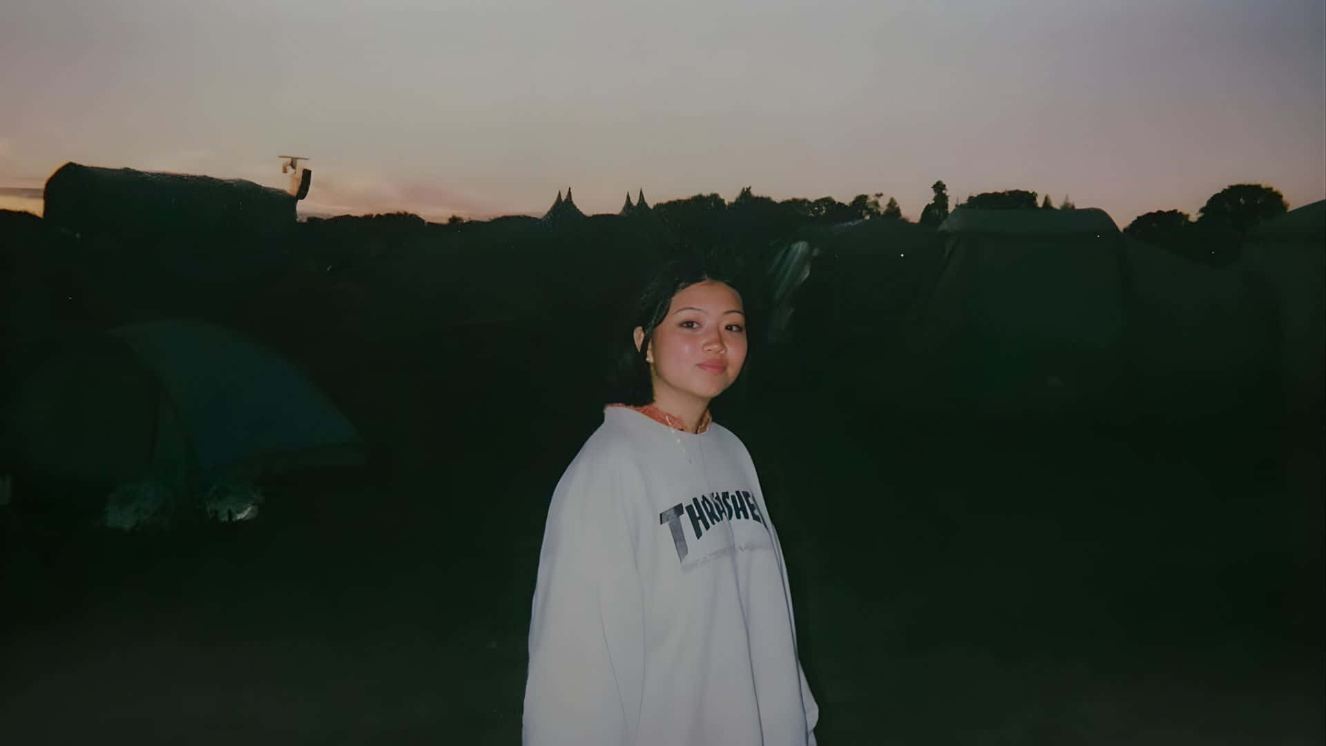  壁紙 — 一位身穿白色衛衣的年輕女子站在黃昏時分燈光昏暗的露營地，身後有帳篷的剪影。