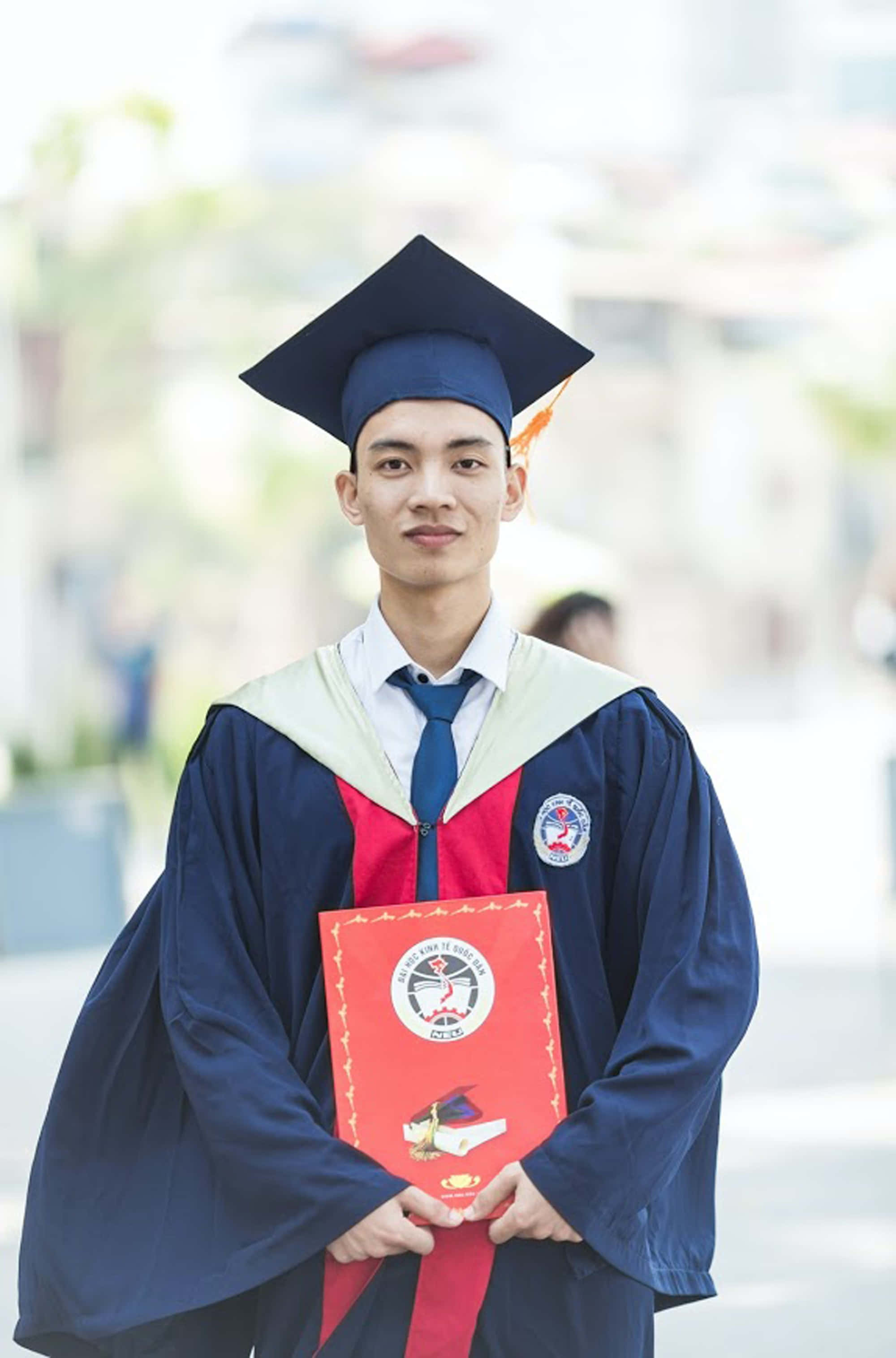 Graduation Ceremony 桌布 壁紙 — 一位身穿海軍藍畢業袍和帽子的年輕畢業生，手持紅色畢業證書套，帶著自豪的笑容，背景是柔和模糊的校園。