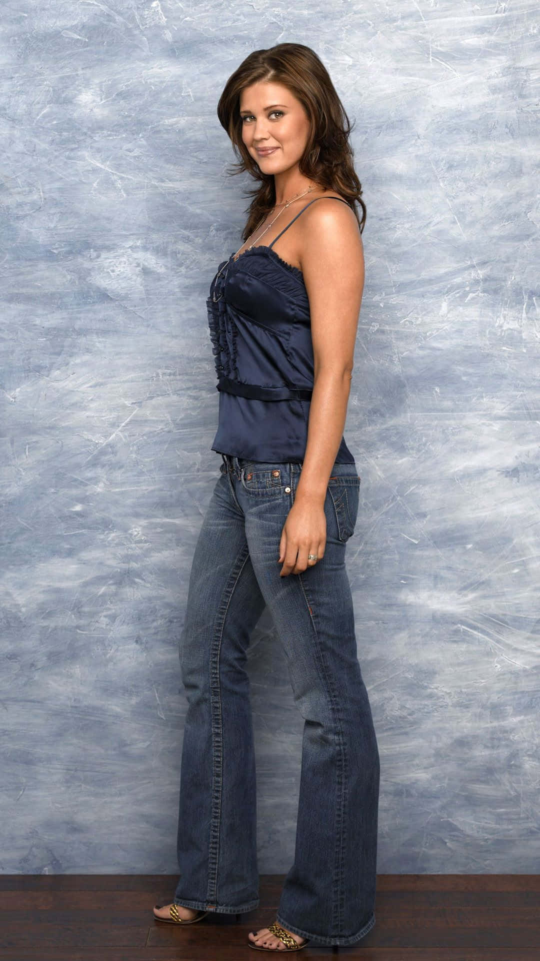 Sarah Lancaster 桌布 壁紙 — 一位身穿海軍藍上衣和喇叭牛仔褲的女性，在帶有紋理的藍灰色攝影棚背景前自信地擺姿勢。