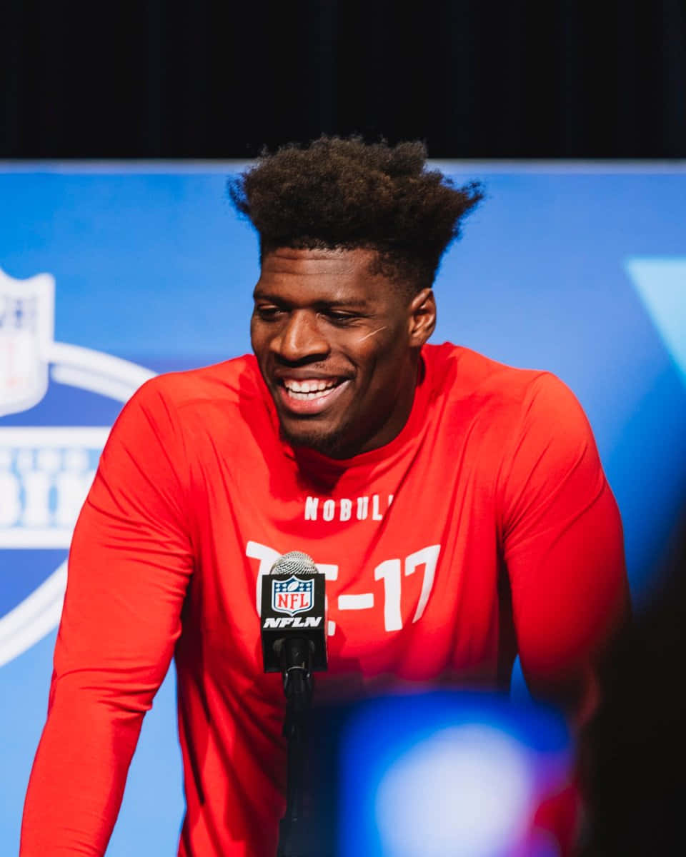  壁紙 — 一位身穿紅色長袖襯衫、面帶微笑的NFL球員在新聞發布會上向麥克風傾身。
