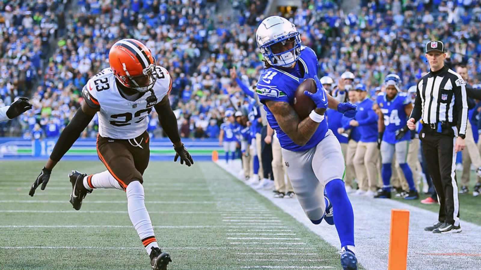 壁紙 — 一位身穿藍色球衣的NFL外接手帶著球衝向達陣區，而一位身穿棕色球衣的防守球員在球場燈光下逼近。