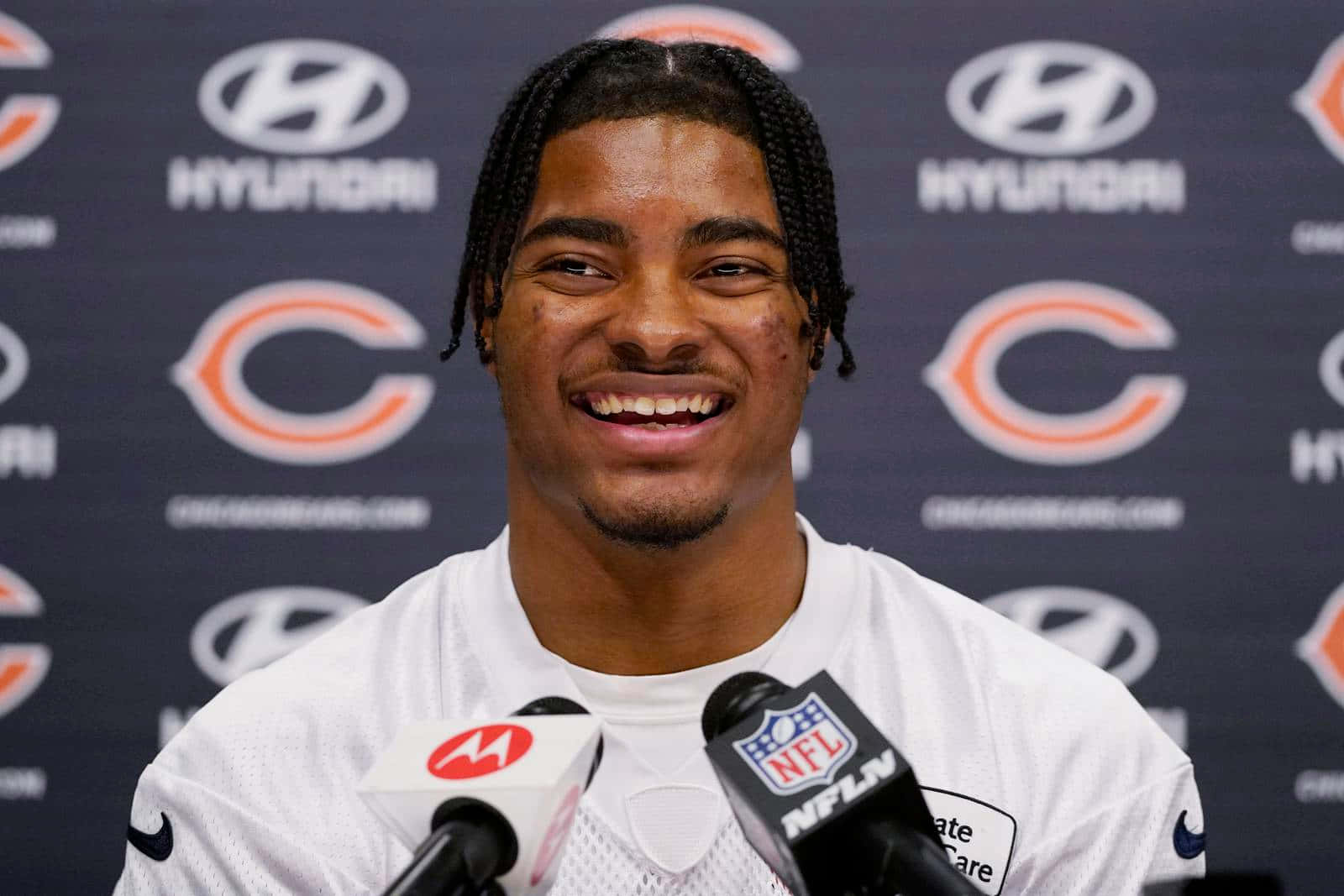  壁紙 — 一位身穿芝加哥熊隊球衣、面帶微笑的NFL球員在品牌背景前的新聞發布會上對著麥克風發言。