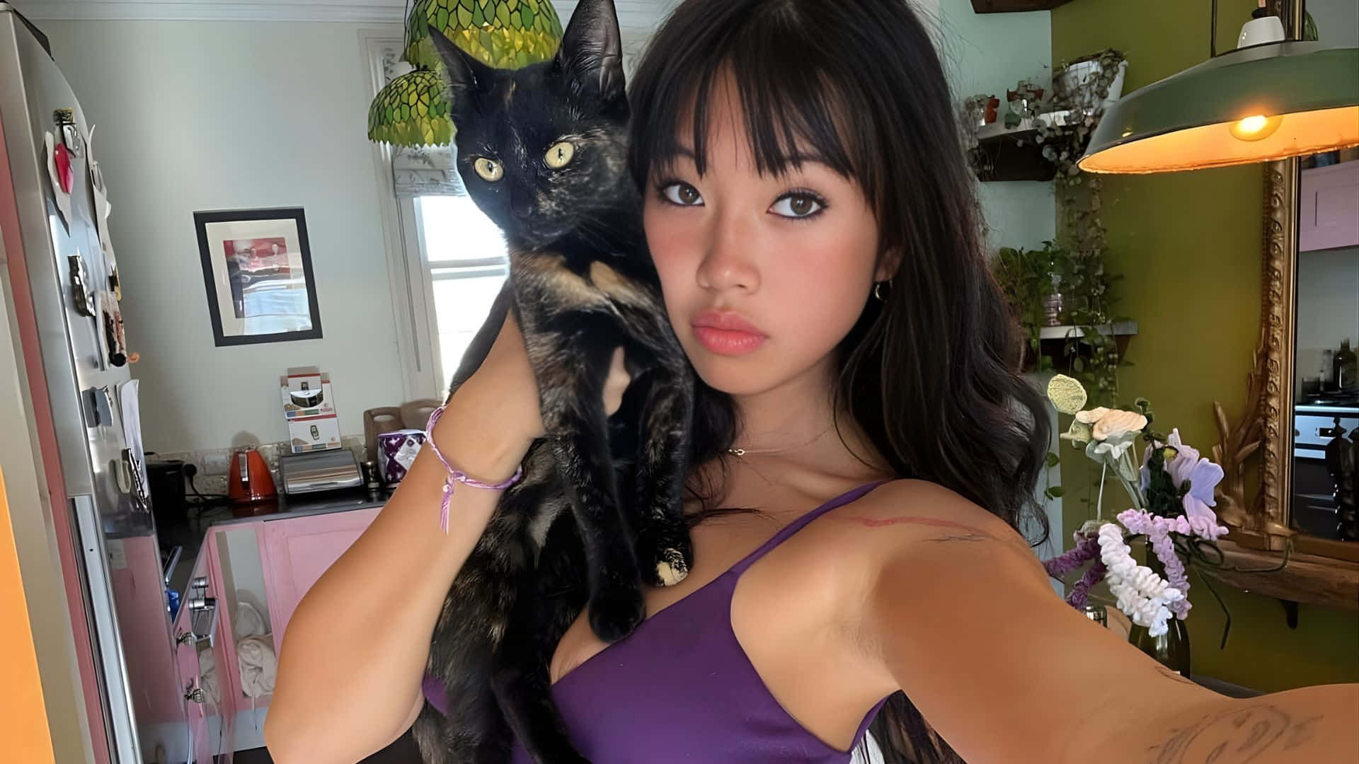  壁紙 — 一位身穿紫色上衣的年輕女子在一個舒適的廚房裡，抱著一隻有著引人注目的黃眼睛的黑貓，廚房裡有綠色吊燈和花卉裝飾。