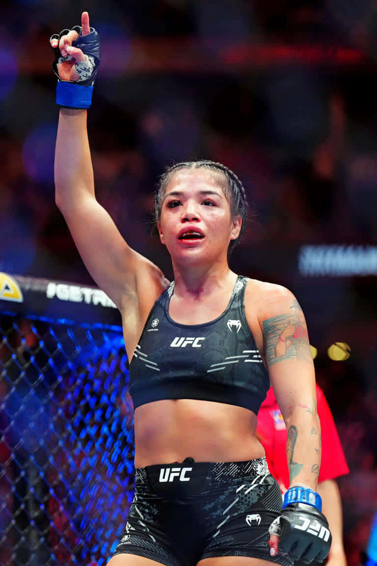  壁紙 — 一位勝利的女性UFC拳手在明亮的場館燈光下，於八角籠內舉起戴著手套的拳頭慶祝勝利。