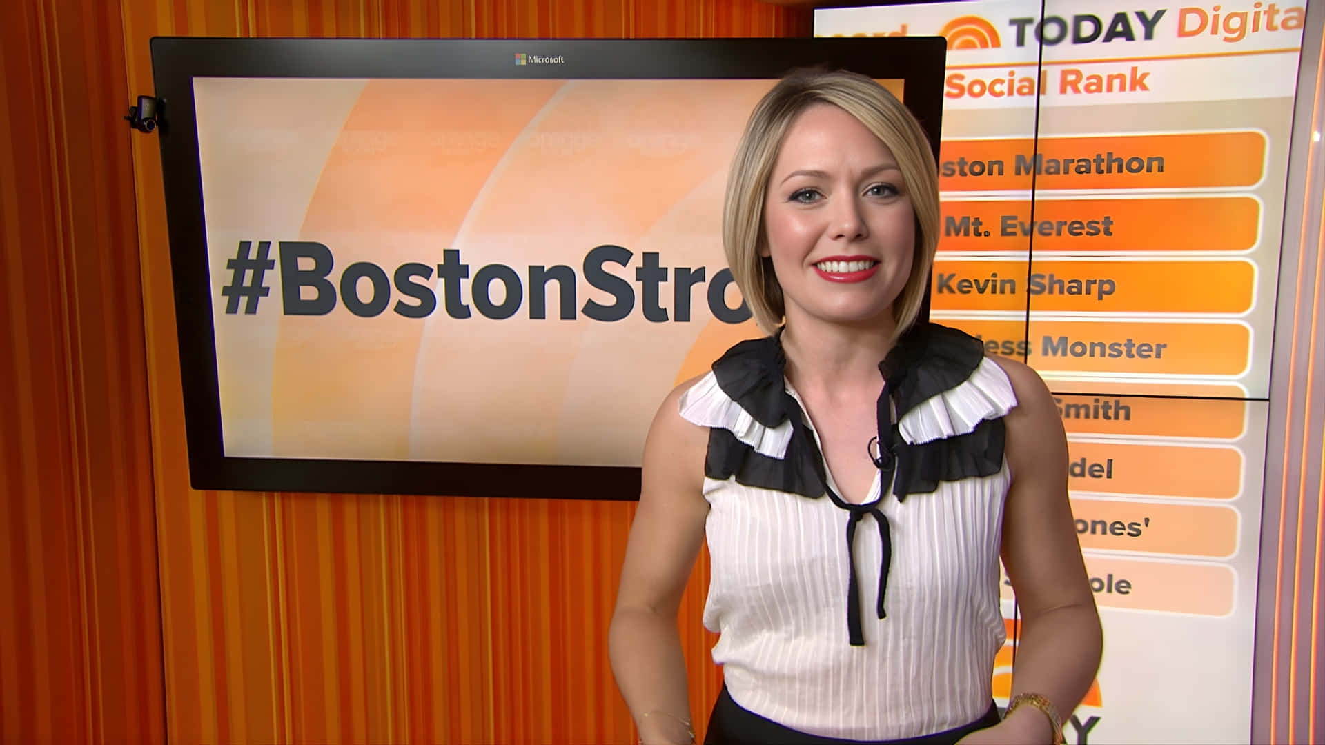  壁紙 — 一位微笑的金髮女主播穿著白色荷葉邊上衣，站在顯示#BostonStrong和社交排名圖形的屏幕前。