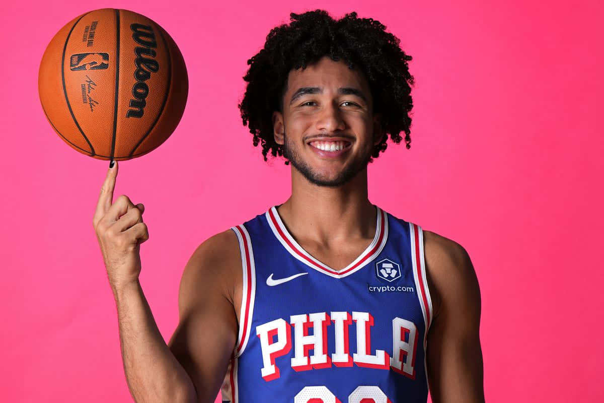  壁紙 — 一位微笑的籃球運動員身穿藍色 'PHILA' 球衣，在充滿活力的粉紅色背景上，用指尖平衡一顆橙色的 Wilson 籃球。