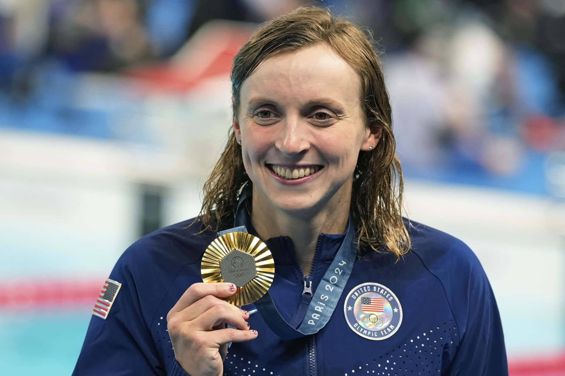  壁紙 — 一位微笑的美國游泳選手在巴黎2024奧運會泳池邊自豪地舉起她的金牌，這是勝利的時刻。