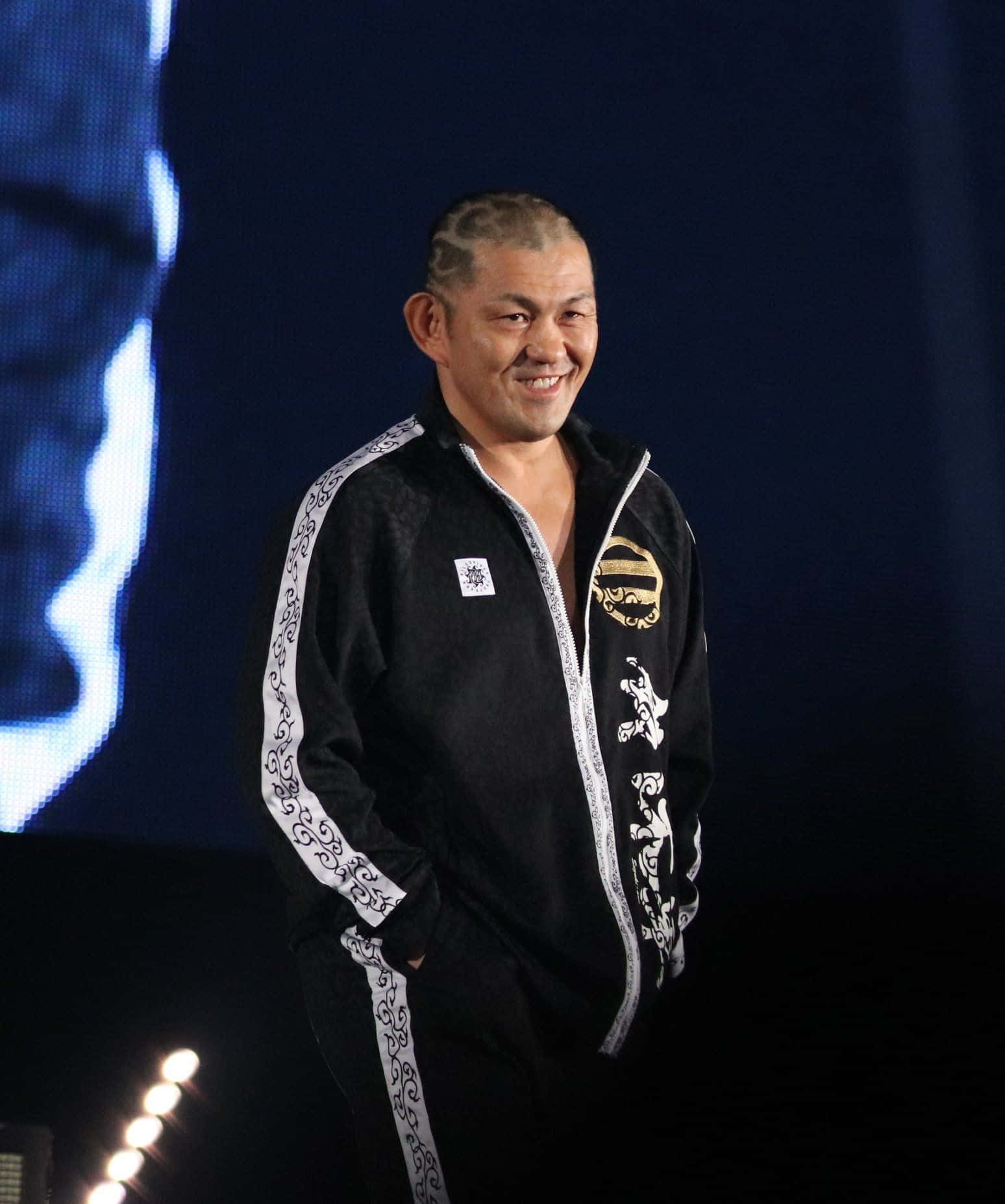  壁紙 — 一位微笑的男子身穿黑白圖案運動服，自信地站在燈光昏暗、帶有藍色照明的舞台上。